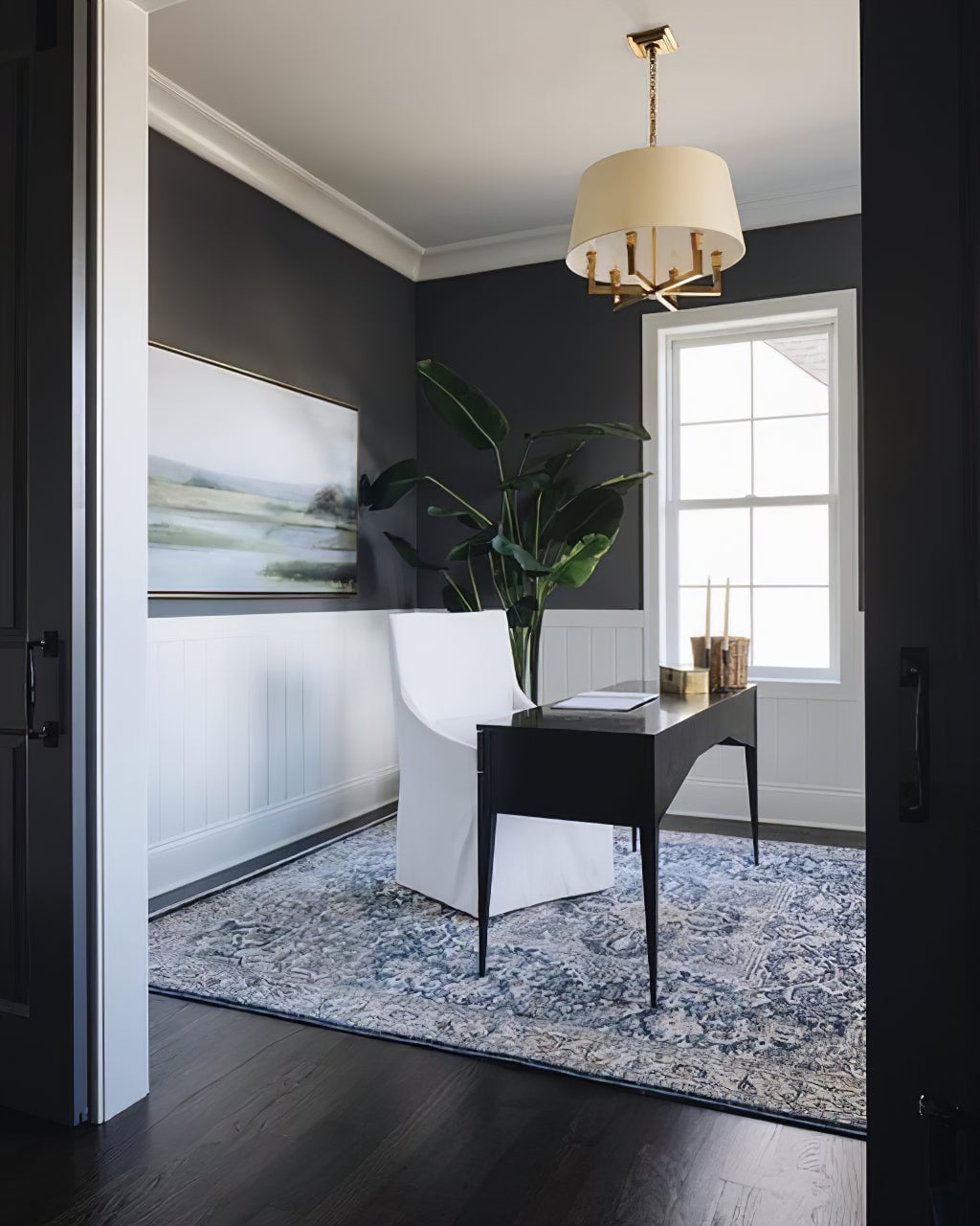Gold-toned shaded chandelier above a dark desk and white chair in a grey room with a large green plant