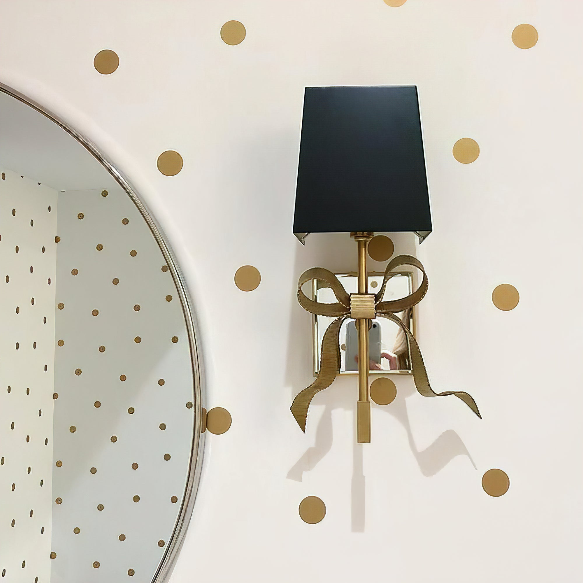 Antique Brass bow wall light with black shade beside a round mirror on gold polka-dot wallpaper