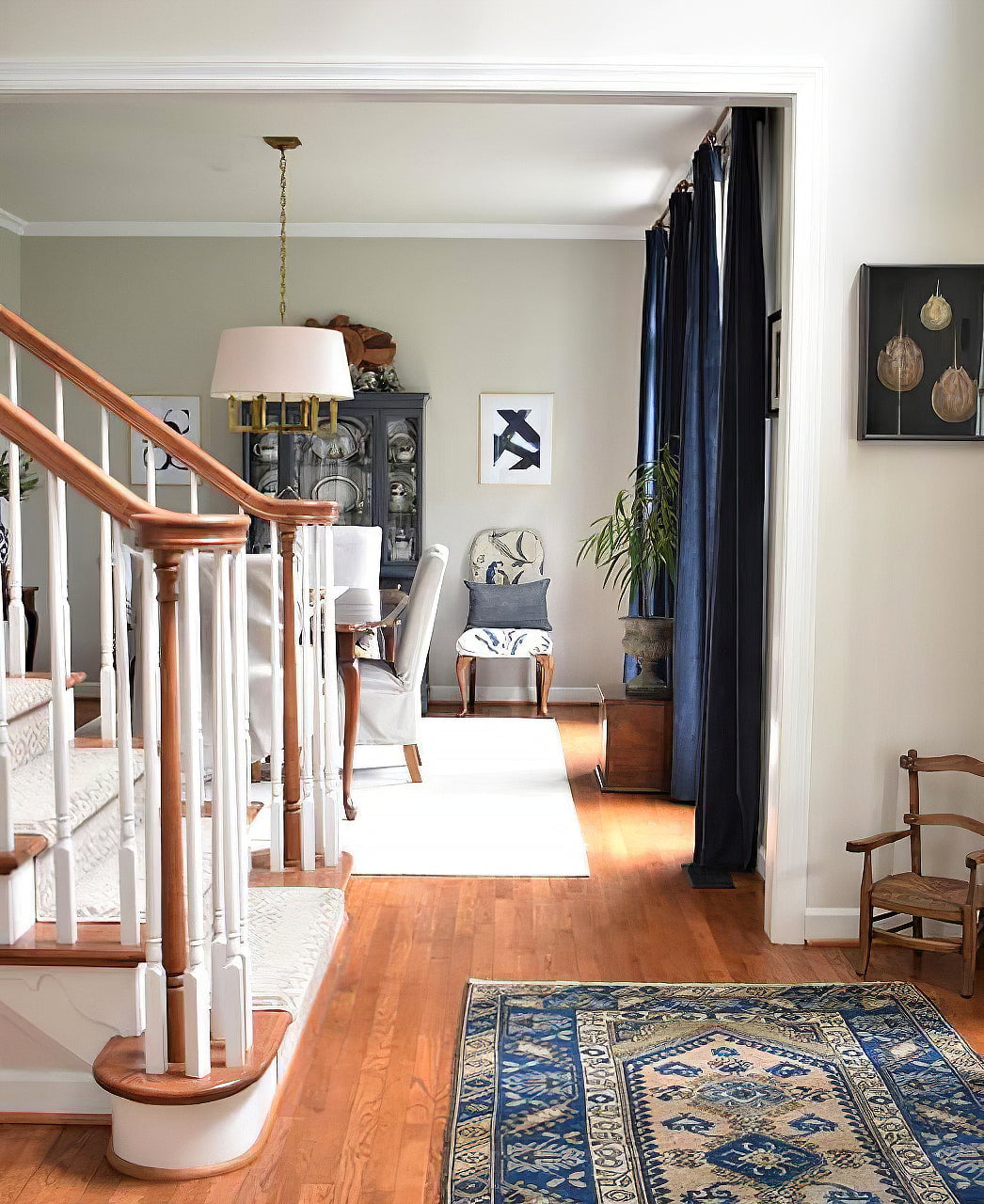 Gold-toned shaded chandelier in a dining room beyond a stair rail, with navy curtains and a china cabinet