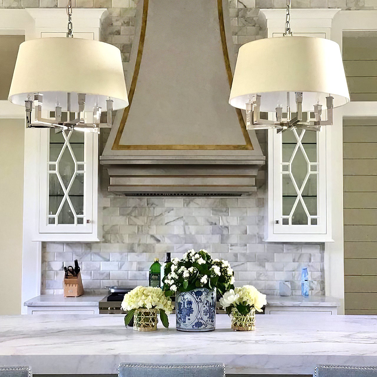Two silver-looking shaded chandeliers over a white island with white flowers in a blue-and-white vase