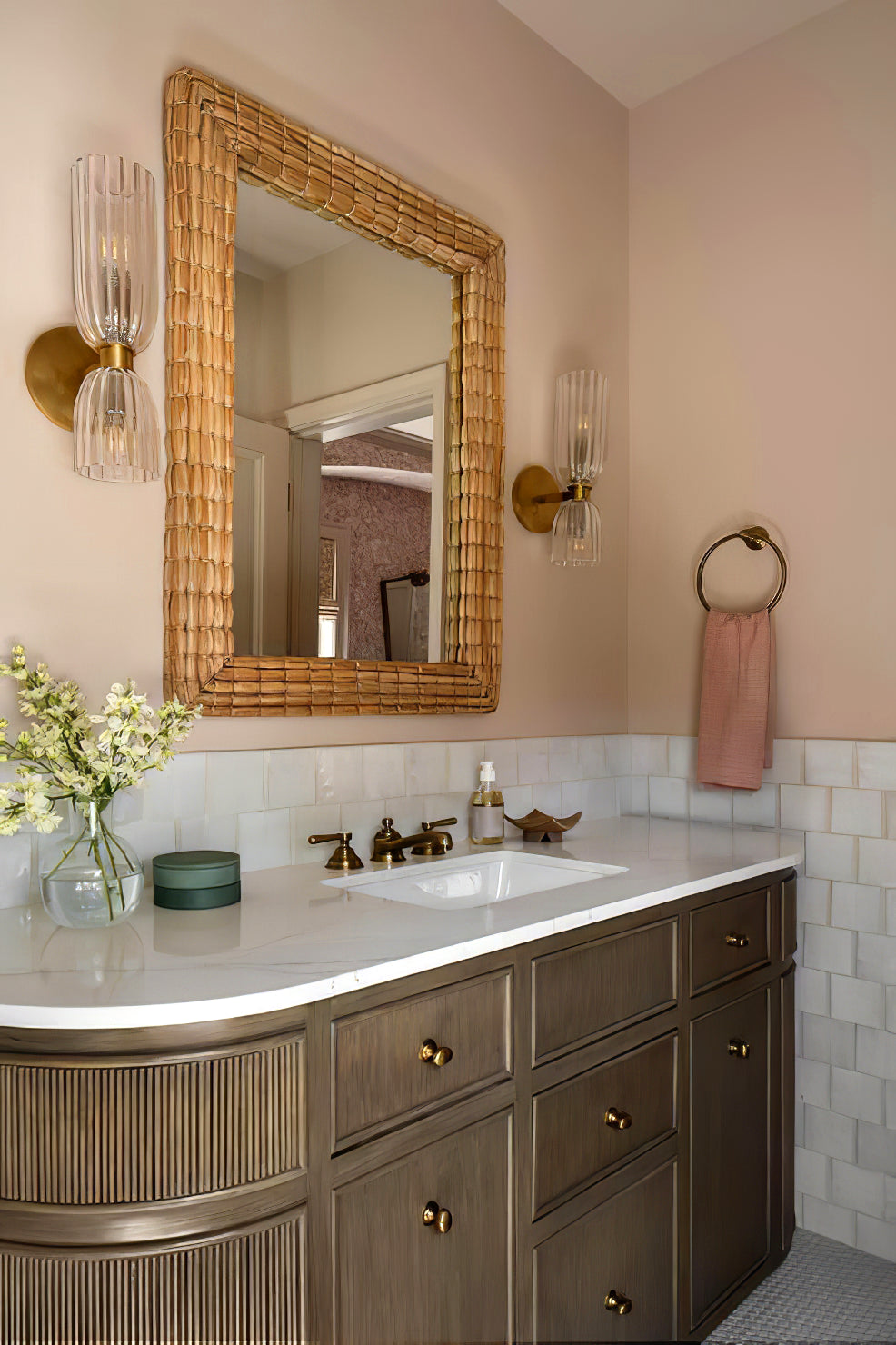 Two brass-tone double glass wall sconces around a woven-look mirror above a curved brown vanity