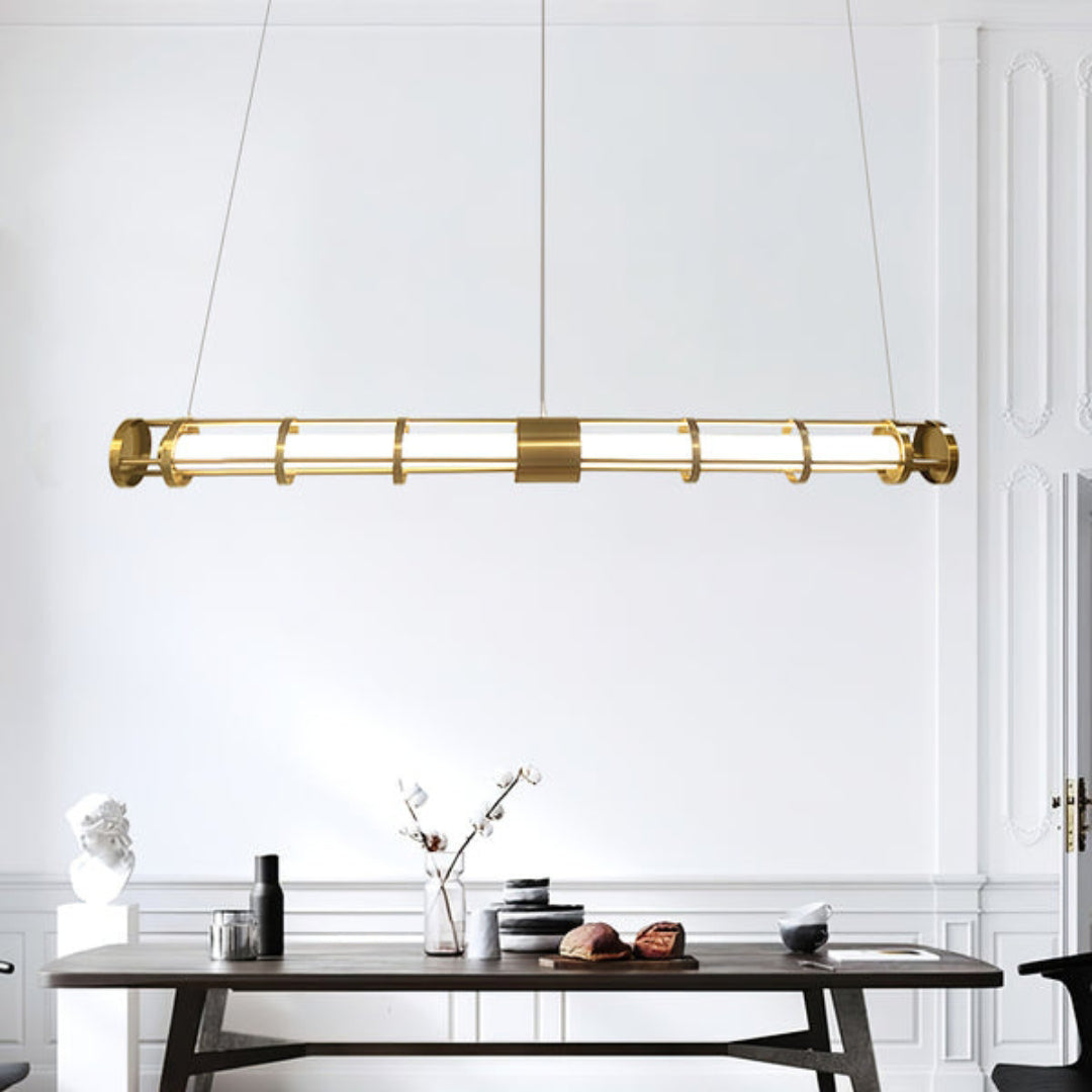 Gold-tone tube pendant above a wooden dining table with bowls, bottles, books and a branch vase