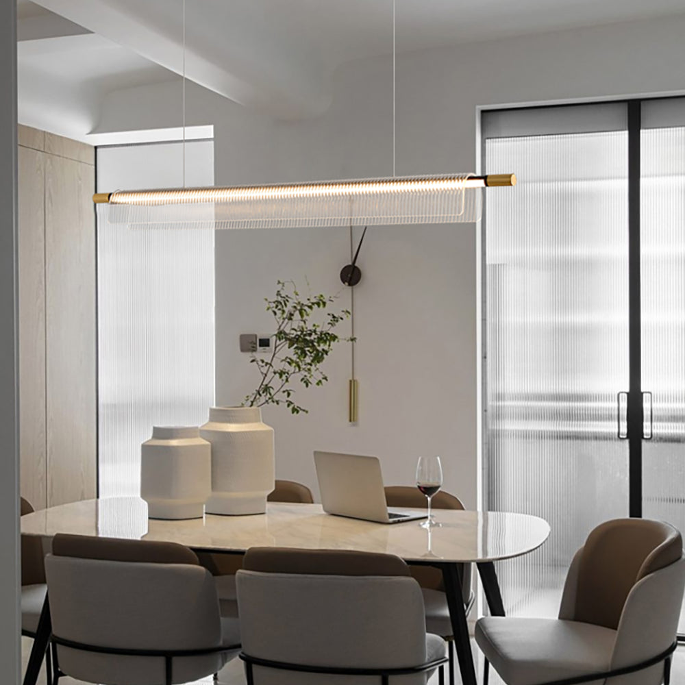 Linear acrylic pendant with gold-tone ends above a pale dining table with white vases and a laptop