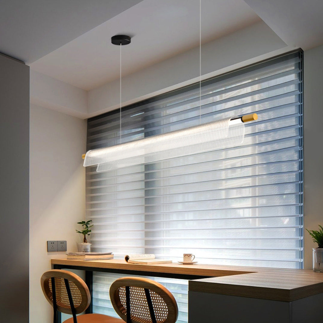 Linear ribbed acrylic pendant with gold-tone ends above a wooden desk beside window blinds and cane-back chairs