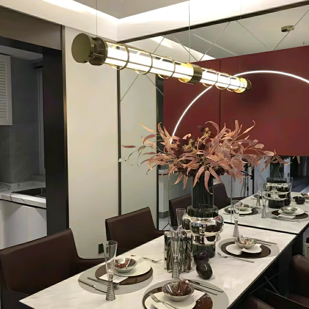 Angled gold-tone tube pendant above a formally set veined table with pink foliage and a mirrored wall
