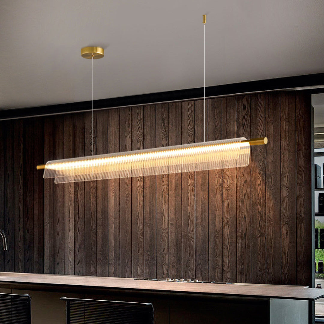 Gold-tone linear pendant with clear ribbed acrylic above a counter against a dark wood-panel wall