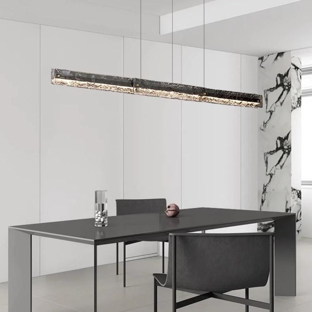 Dark-framed linear glass pendant above a narrow black table near a veined stone-pattern wall