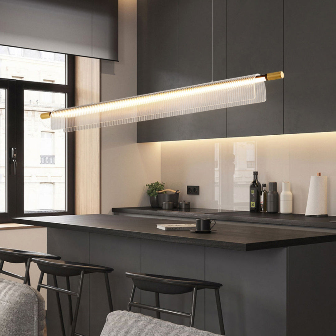 Clear ribbed linear pendant with gold-tone ends above a dark kitchen island, stools and cabinets