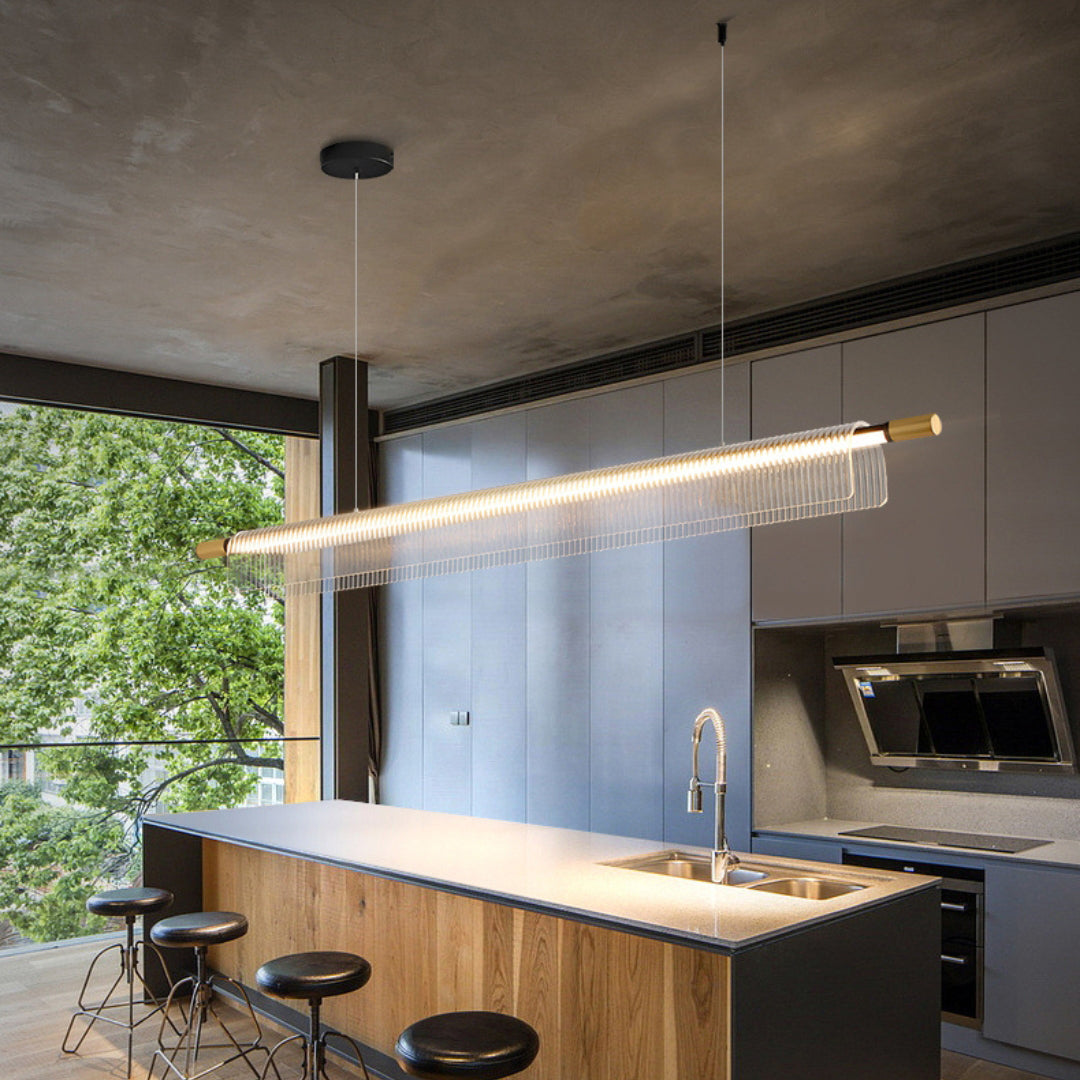 Long illuminated ribbed acrylic pendant above a wooden kitchen island beside a large leafy window