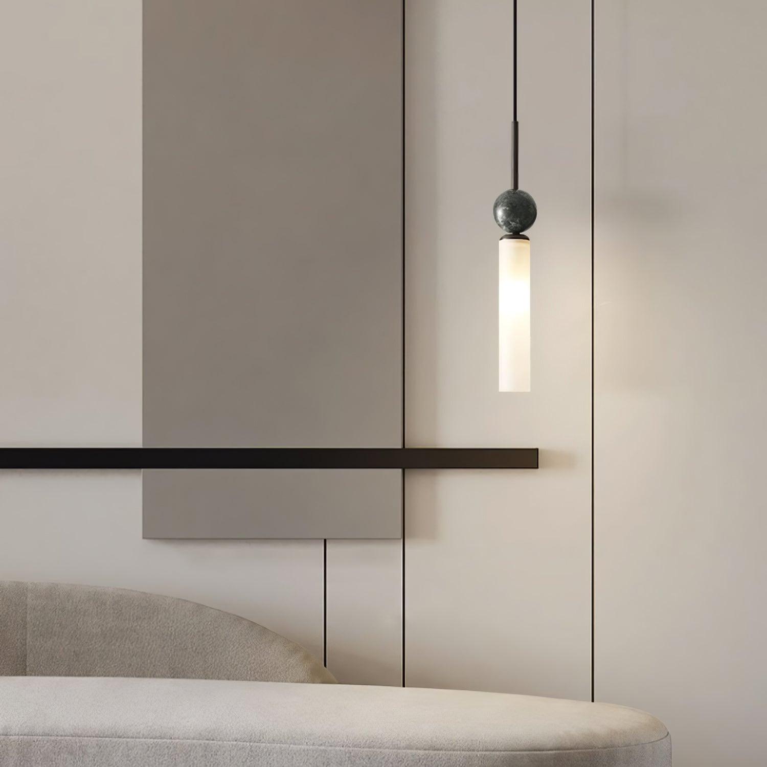 Black-stem pendant with veined dark marble sphere and lit white tube above pale curved sofa and paneled wall