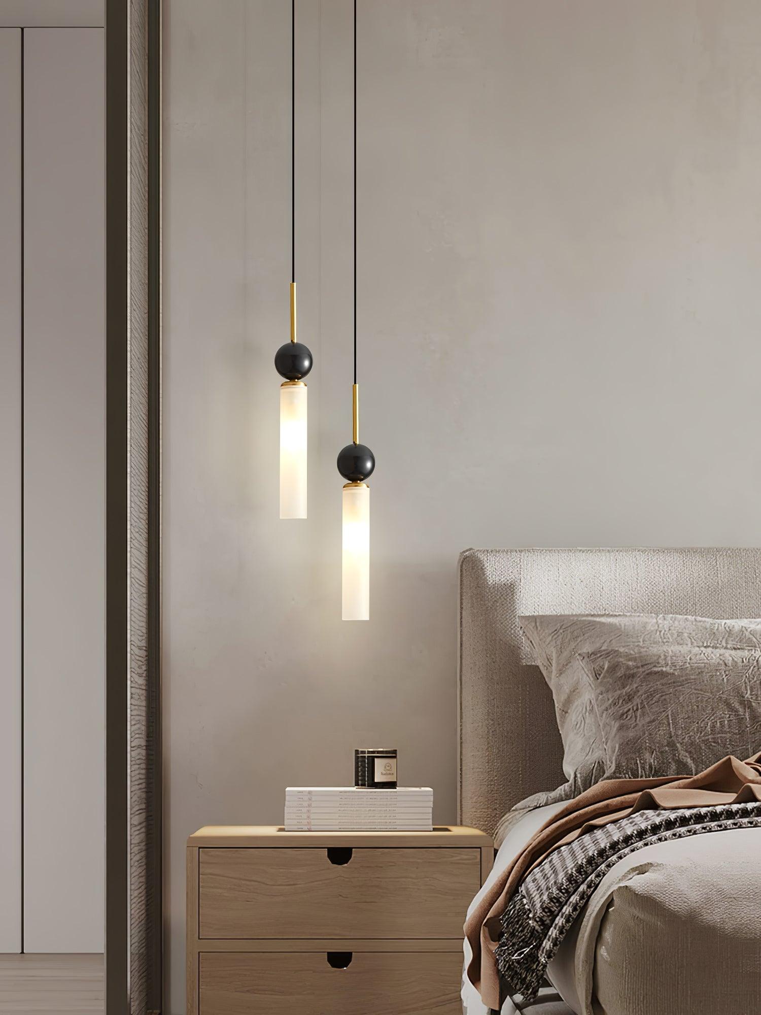 Wide bedside view of two staggered brass-stem pendants above wood nightstand, with books, candle and upholstered headboard
