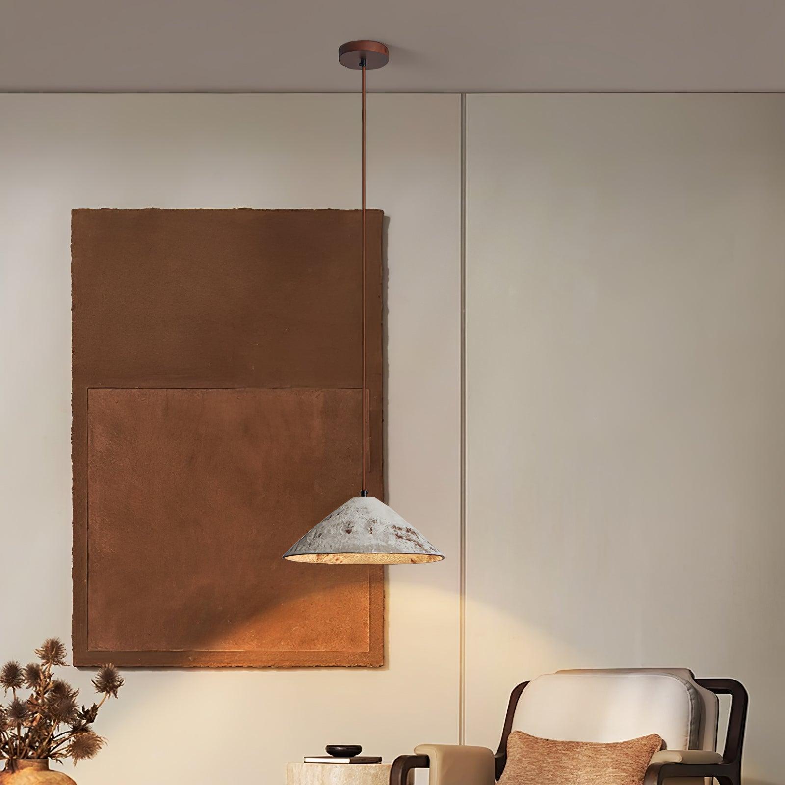 White ceramic cone pendant on a brown cord beside a cream armchair and large brown wall art, with dried flowers at left