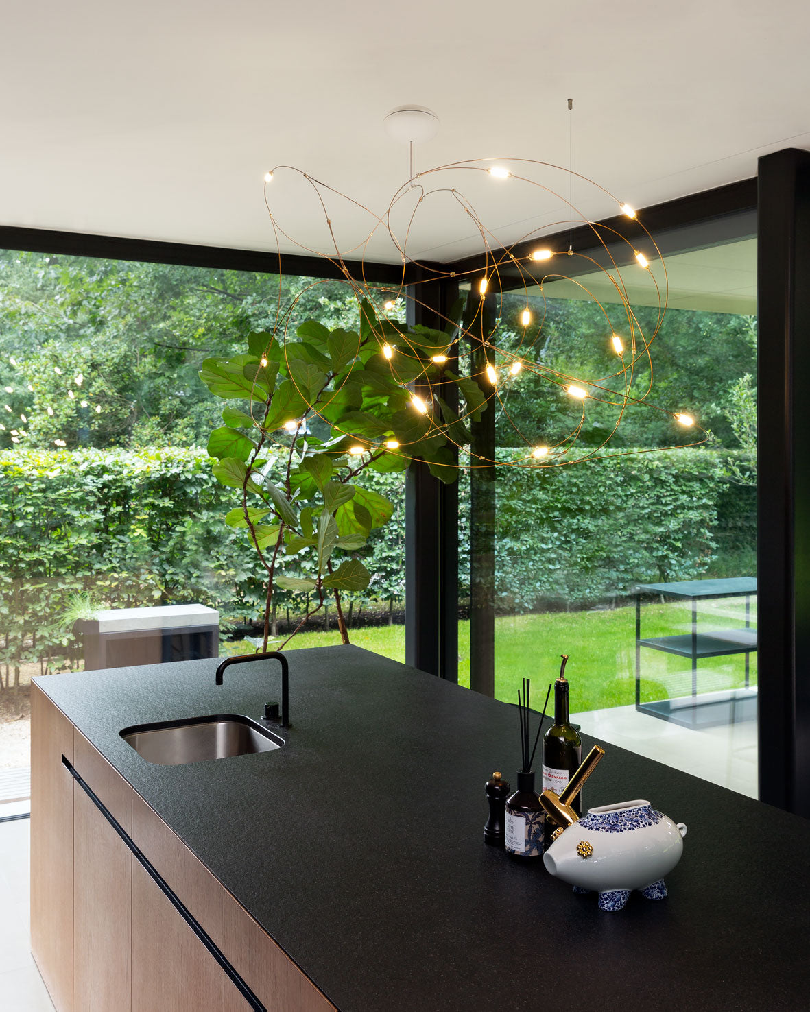 Gold-tone looping-wire pendant lit above a dark kitchen island near a plant and large windows