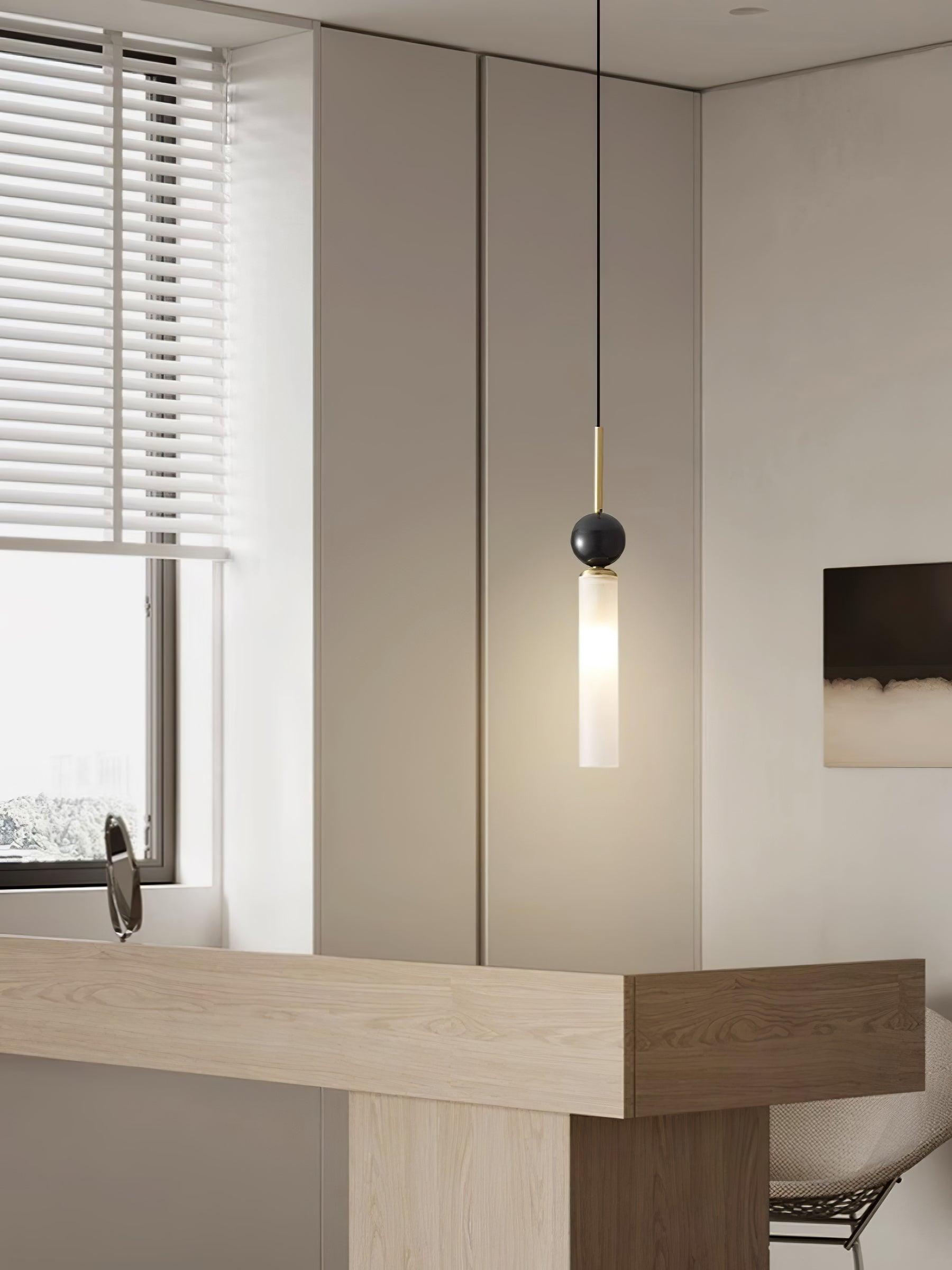 Brass-stem dark-sphere pendant above wood desk beside window blinds, tabletop mirror, mesh chair and wall art