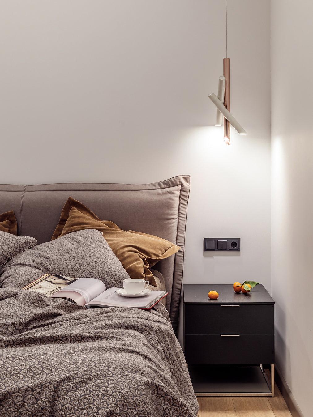 Lit white and rose-gold Tubes pendant above a dark bedside cabinet beside an upholstered bed