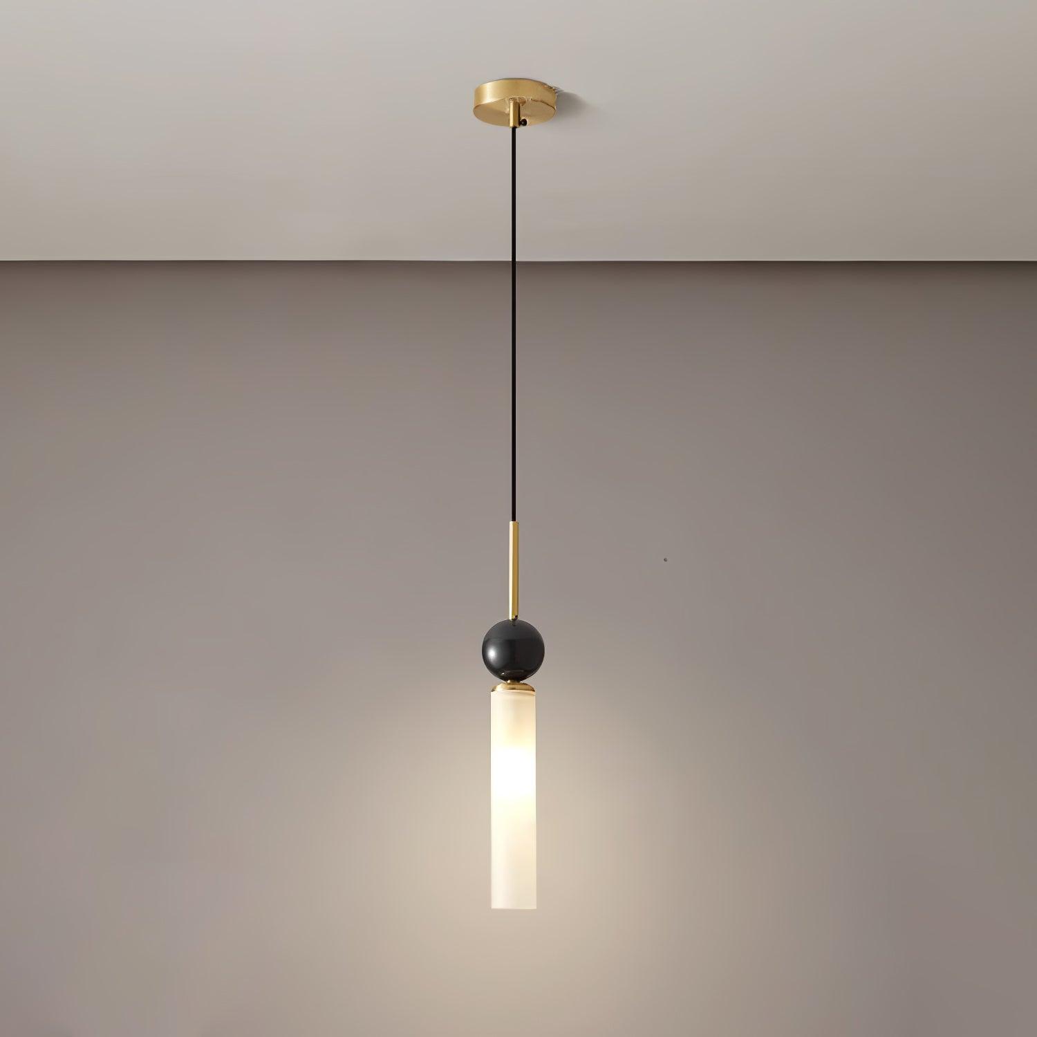 Brass-tone single pendant with small round canopy and dark sphere, glowing white tube and warm light pool on gray wall