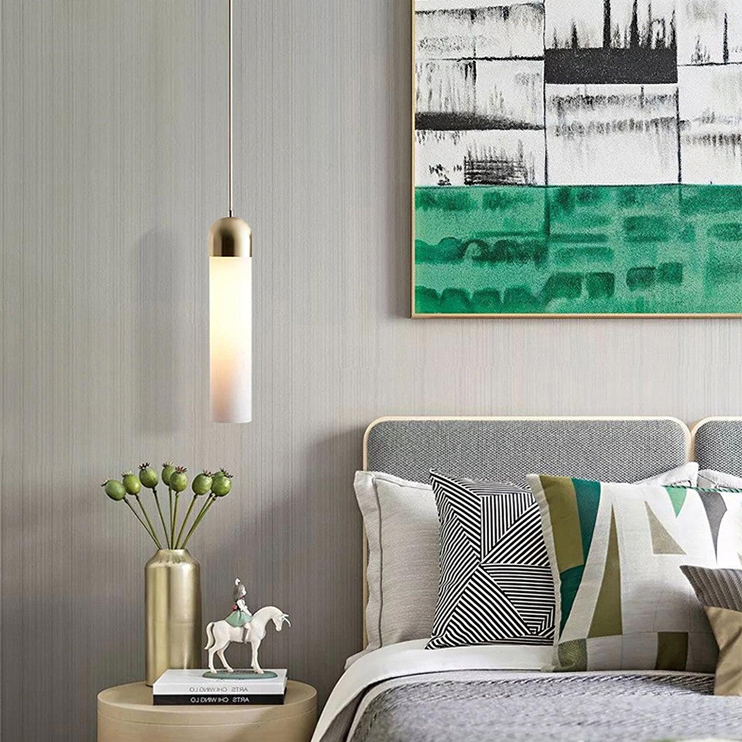 Milk white cylindrical pendant with gold-tone cap beside a bed and round nightstand