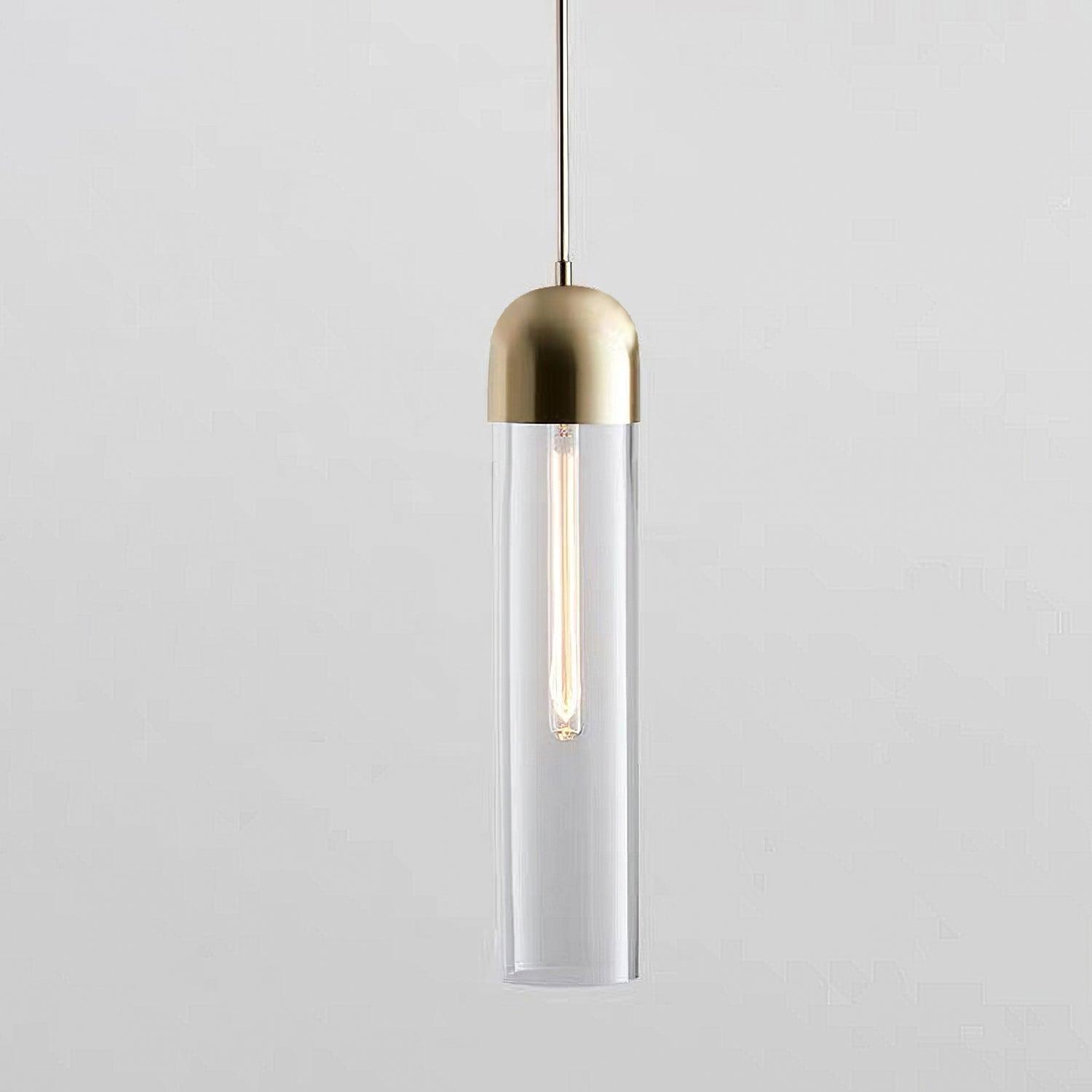 Clear glass cylinder pendant with gold-tone cap and lit elongated filament bulb on pale grey
