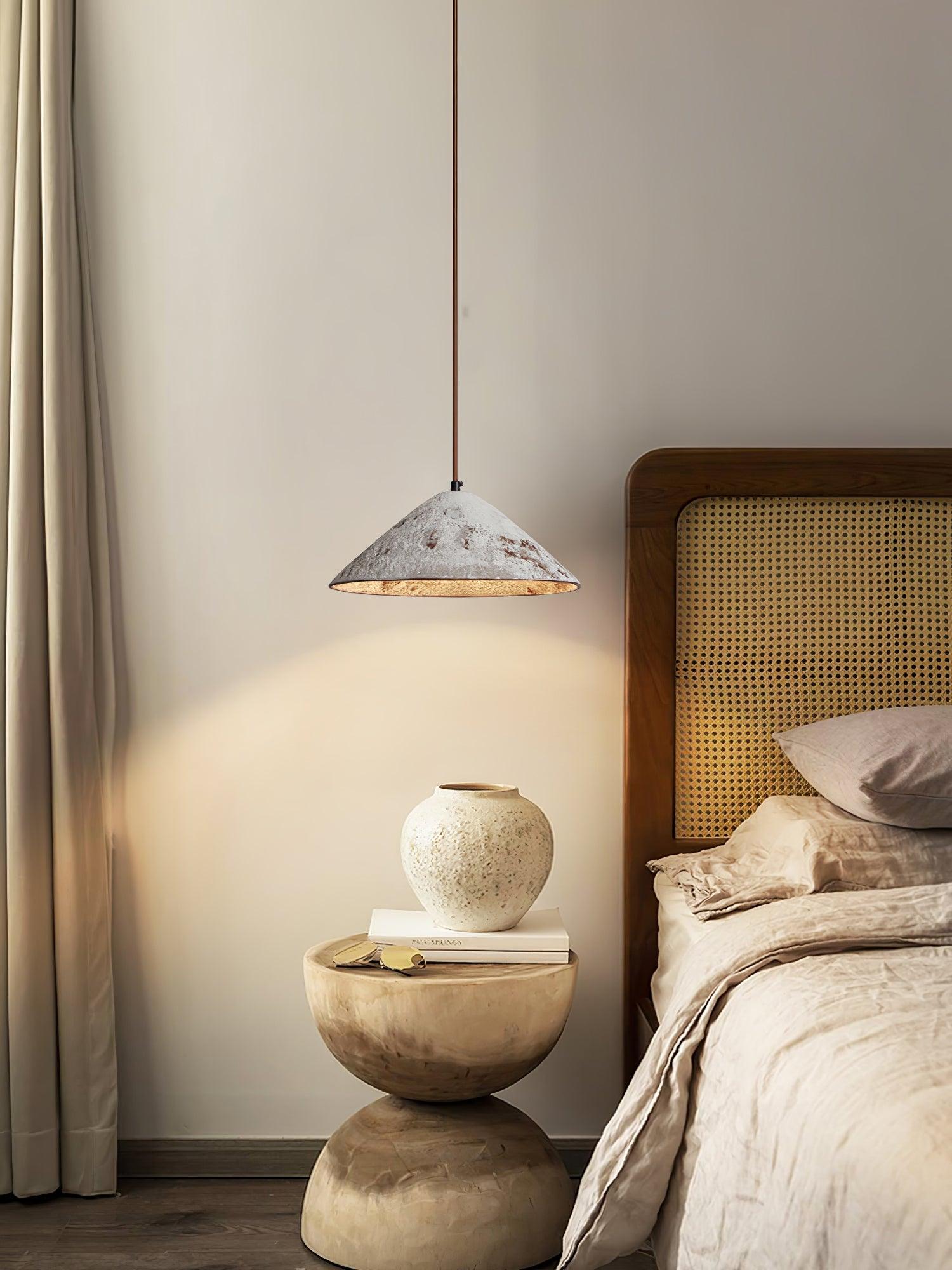 White cone pendant beside a woven headboard, above a round stone bedside table with a white vase, books and sunglasses