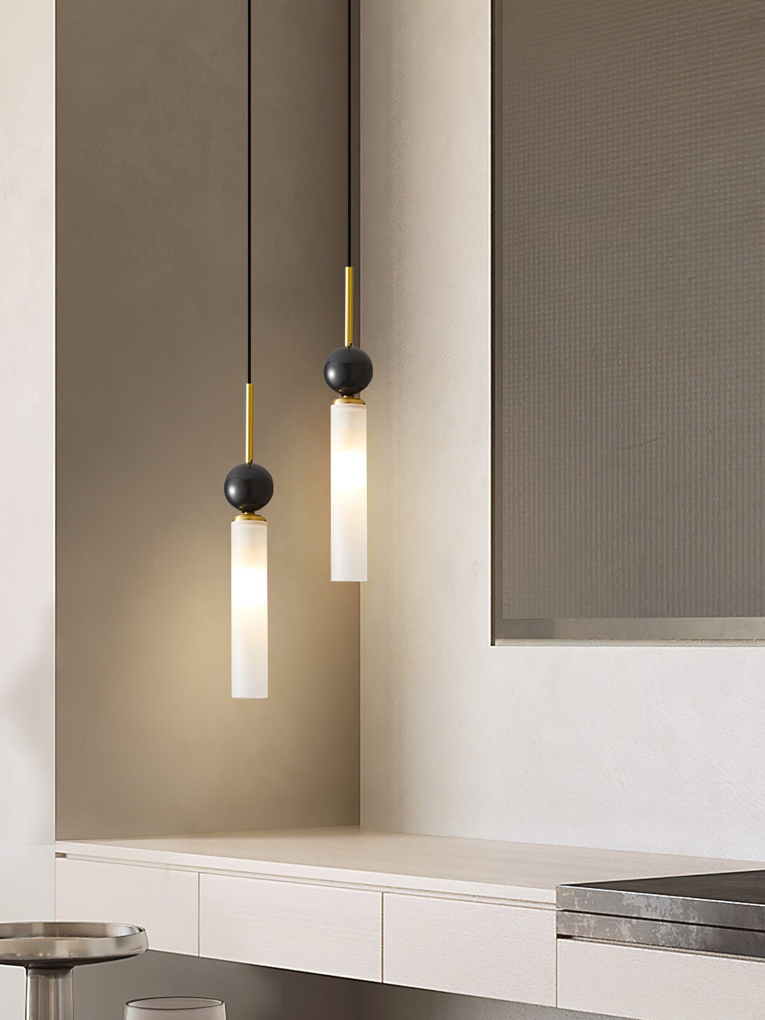 Pair of staggered brass-stem pendants beside abstract artwork and pale cabinet, with a metal tray and glass in front