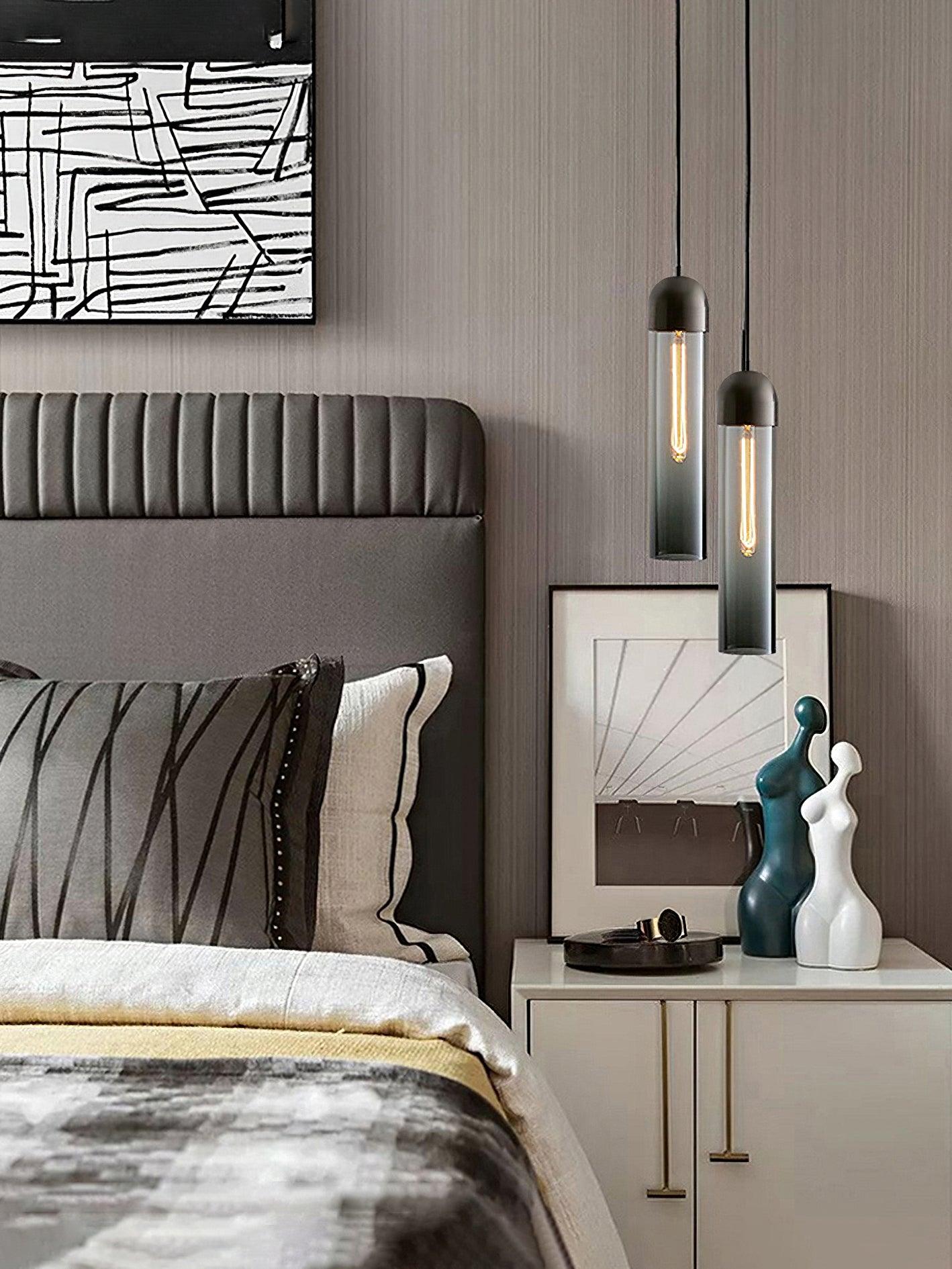 Two smoky glass cylinder pendants at different heights beside a grey upholstered bed