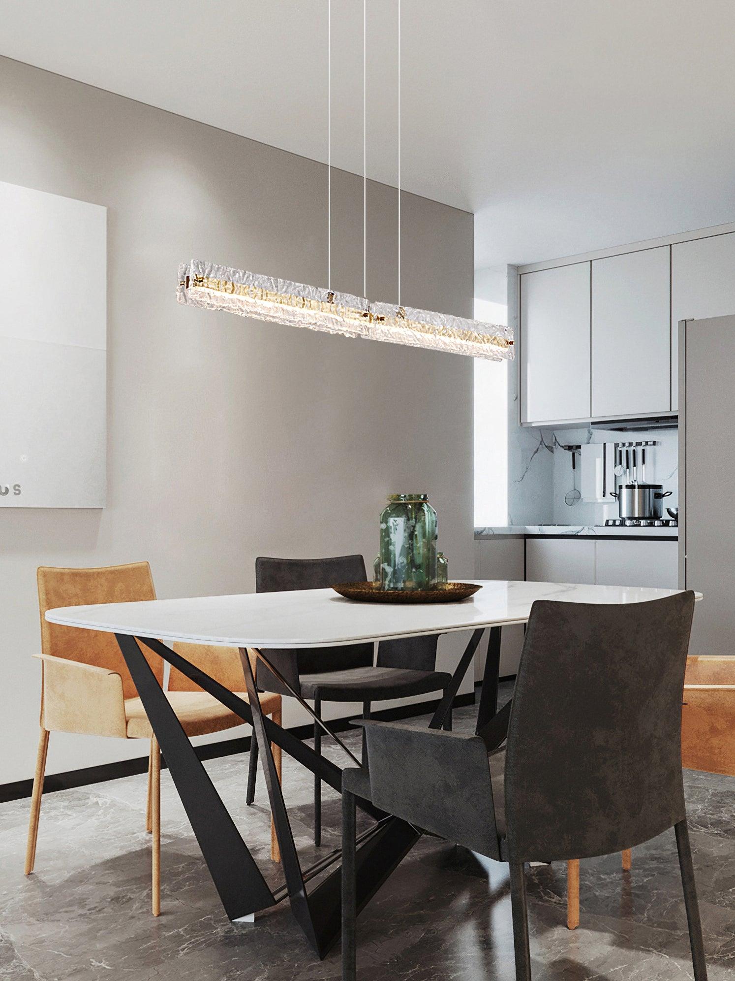 Clear rectangular linear pendant with brass-tone strip above a pale table and green vase near kitchen cabinets