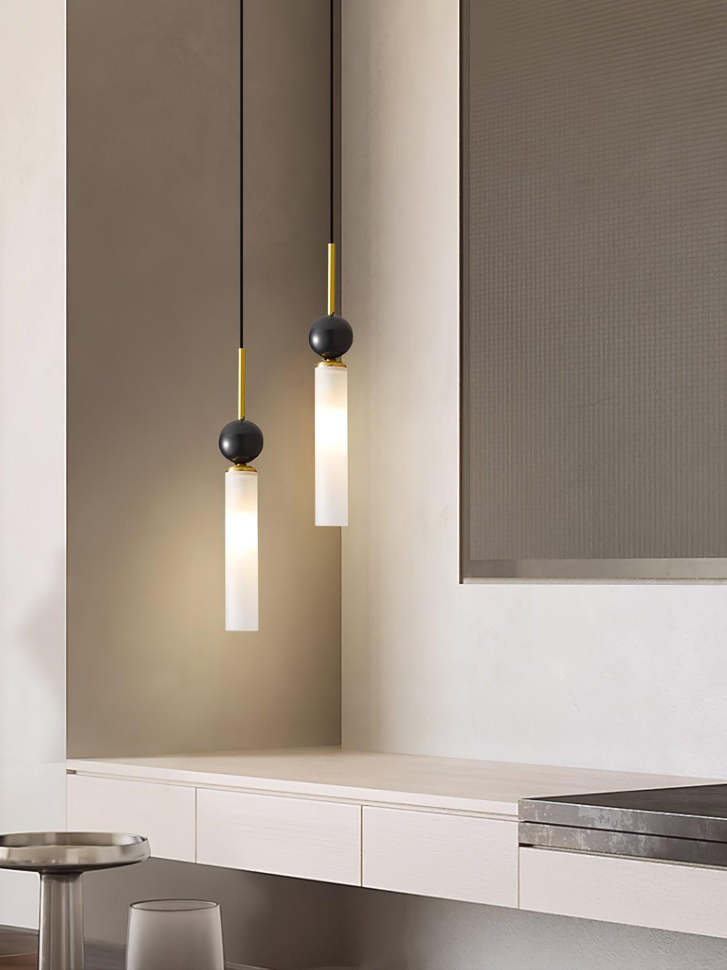Two brass-stem dark-sphere pendants with white lit tubes over pale cabinet beside artwork, tray and glass