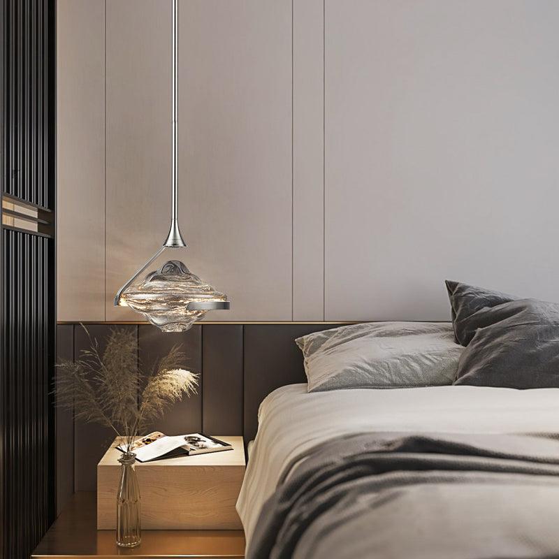 Chrome-looking rippled-glass pendant beside a grey bed, with wooden table, open book and dried stems