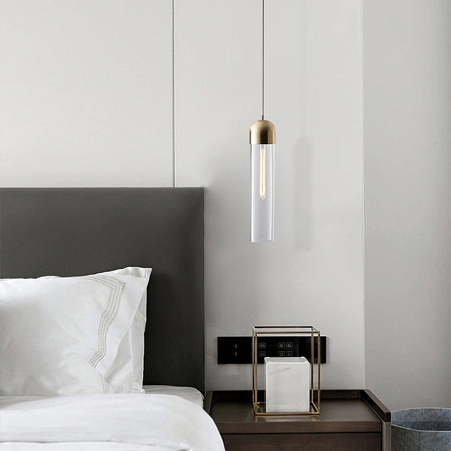 Clear glass cylinder pendant with gold-tone cap beside a bed and bedside table