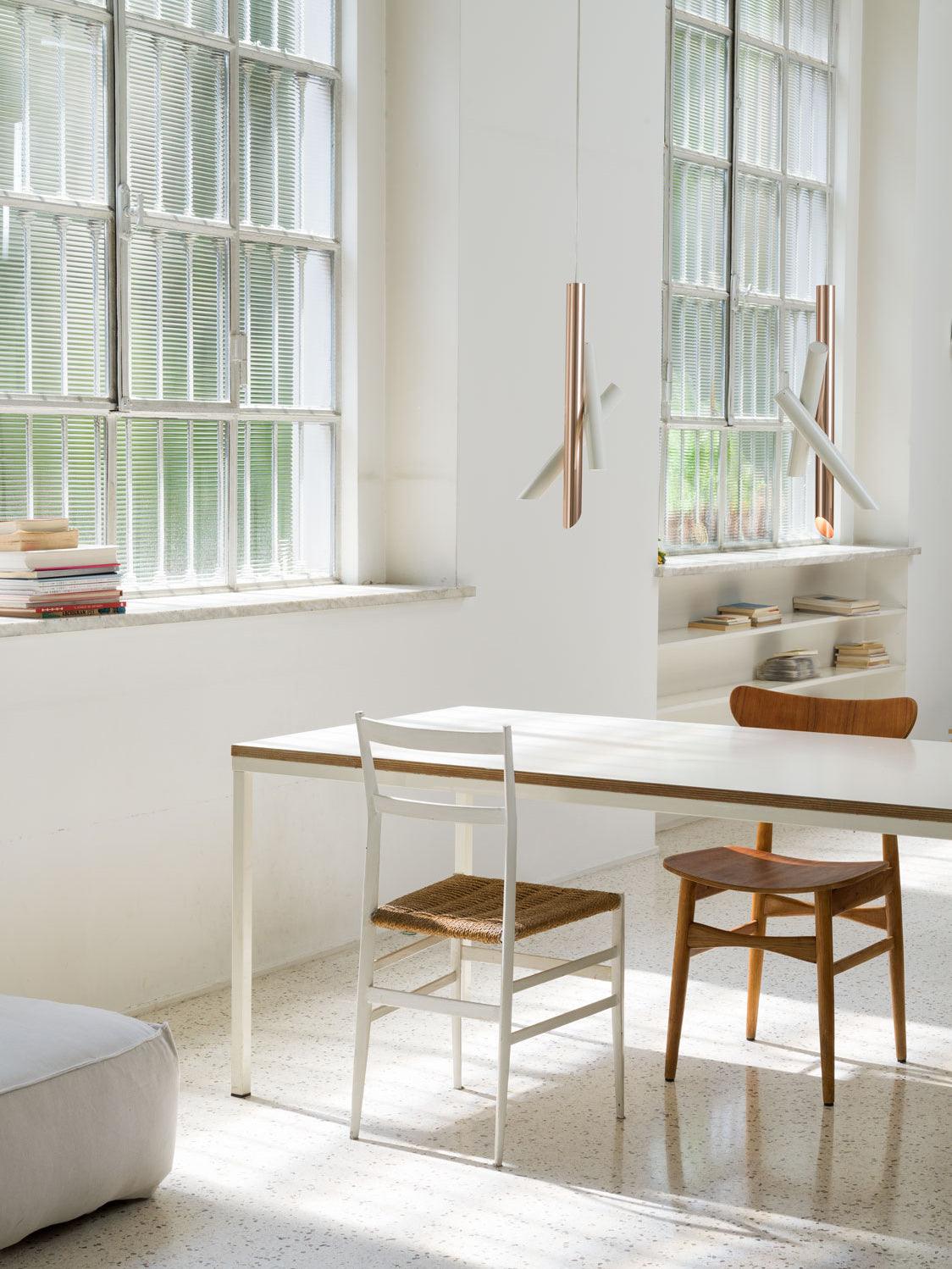 White and rose-gold Tubes pendants above a table beside tall windows and shelves of books