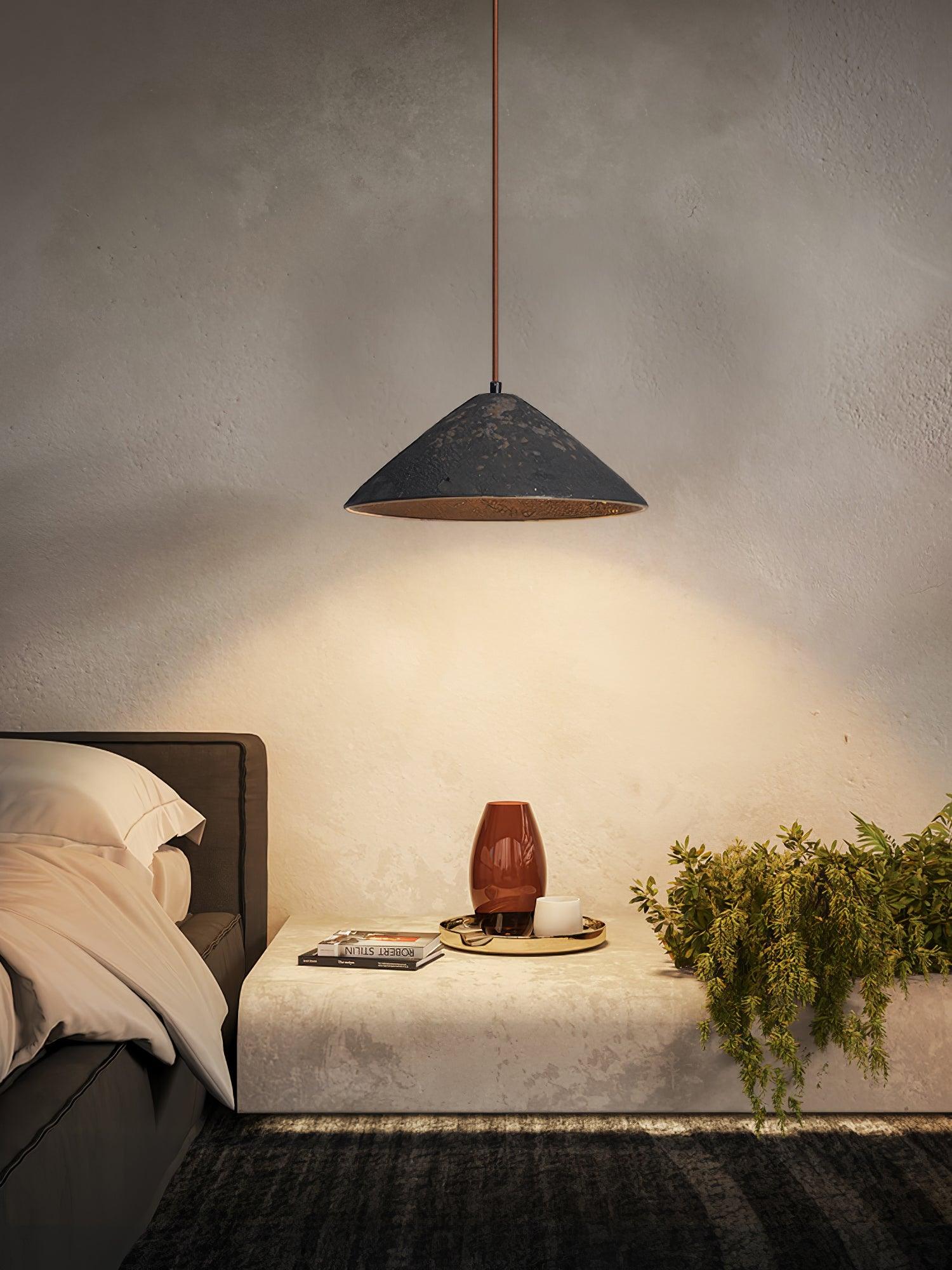 Black ceramic cone pendant over a stone bedside bench with a red vase, white cup, books and green trailing plant
