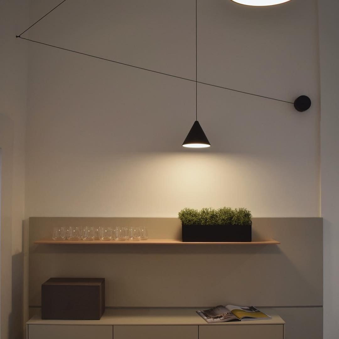 Lit black conical pendant over a wood shelf with glasses and plants, with black cable routed across the wall