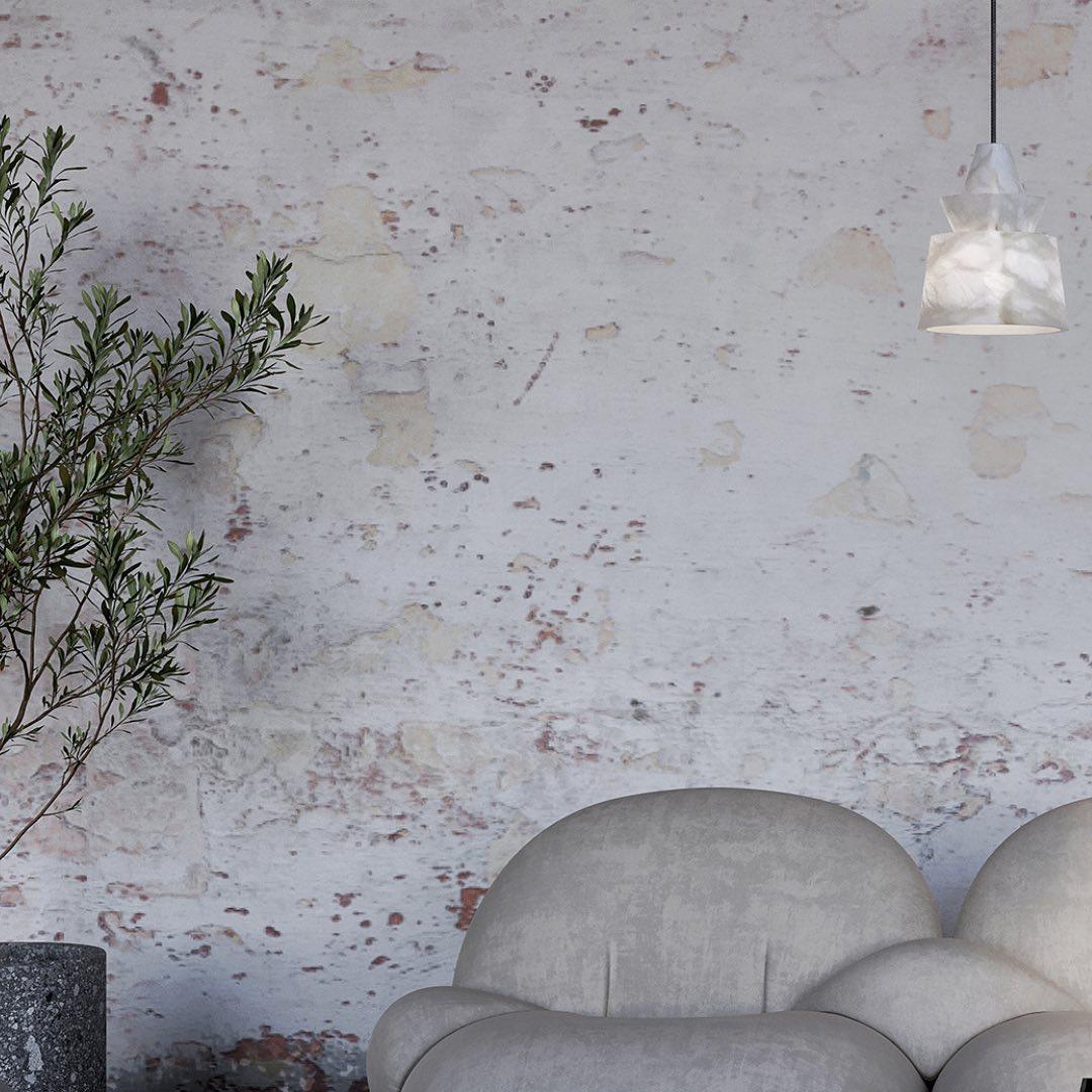 Single glowing broad-bottom stone pendant above rounded sofa arm, beside leafy plant and distressed pale wall.