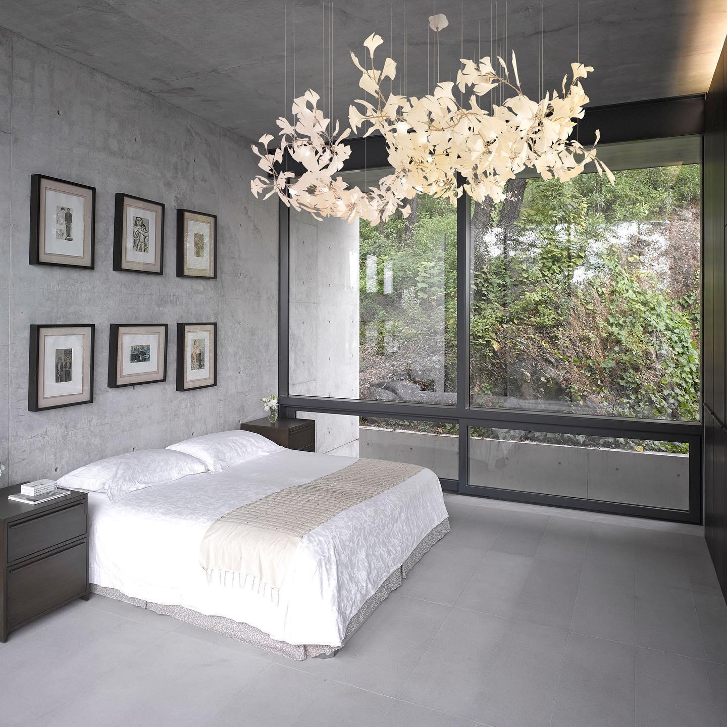White branching leaf chandelier above a white bed in a grey concrete room with a garden-facing window