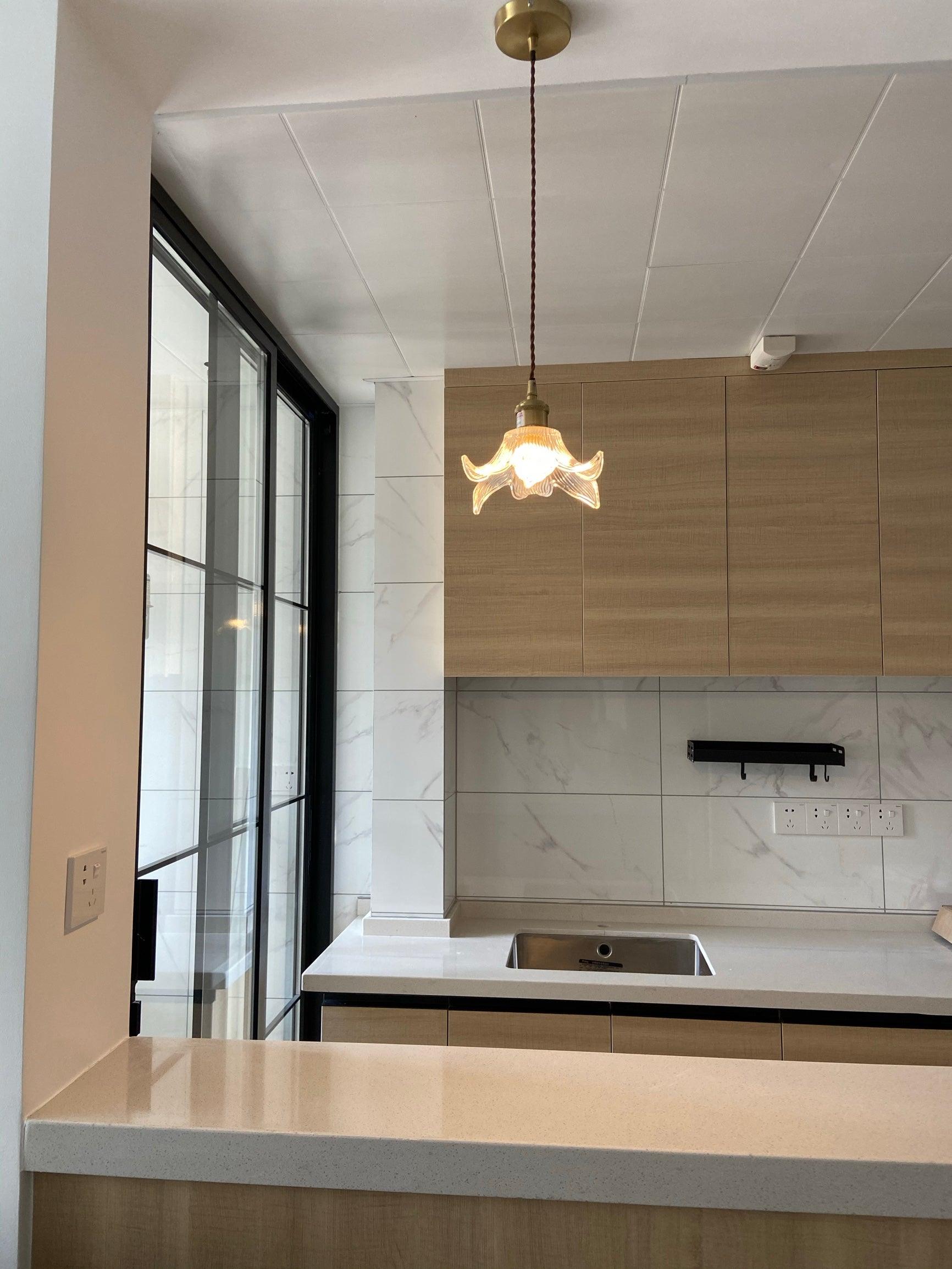 Single lit petal pendant and round brass-toned canopy in a kitchen with wood cupboards, white counter, sink and black-framed glass door.