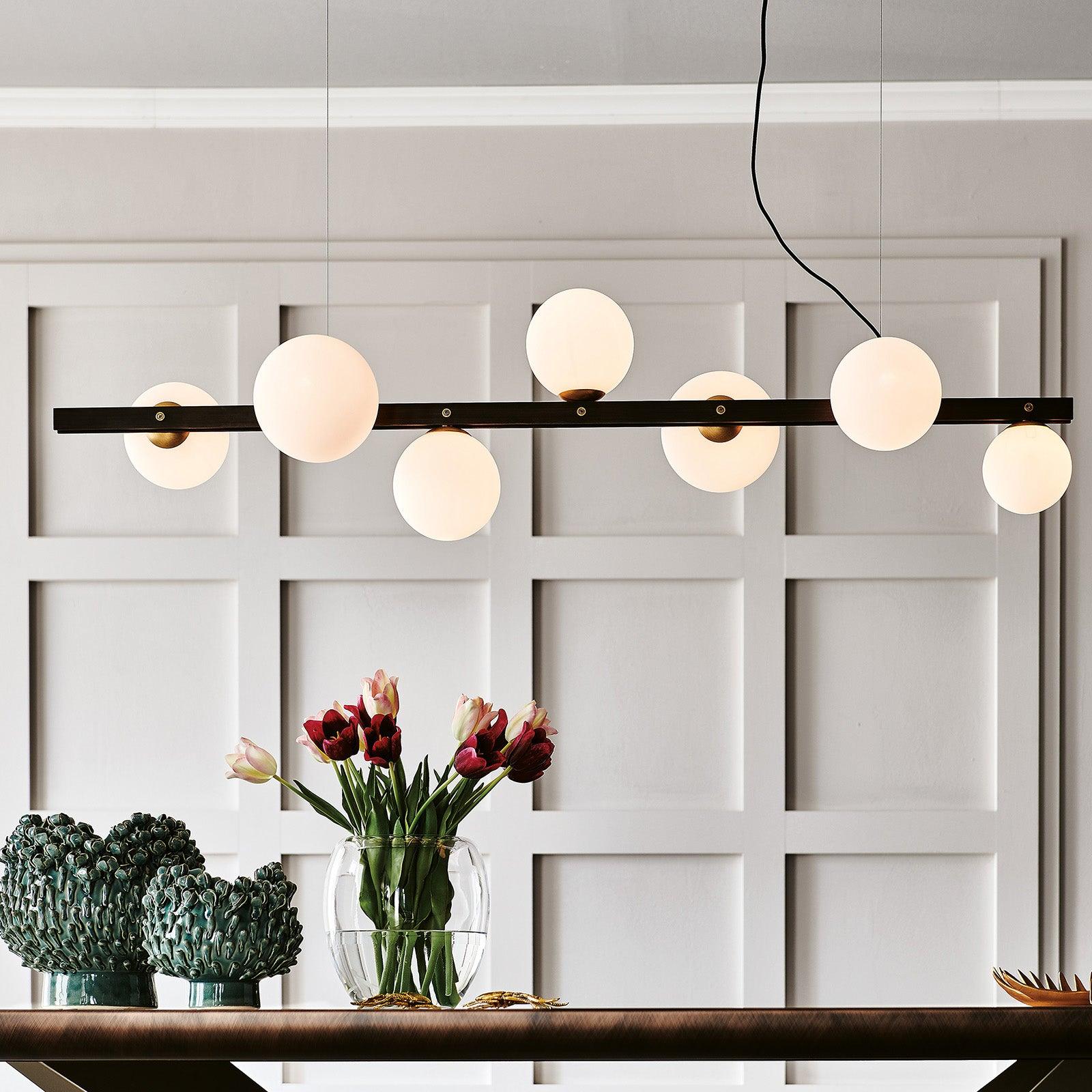 Seven glowing globes along a black bar above tulips and green textured bowls, against white panelling with fine suspension lines.