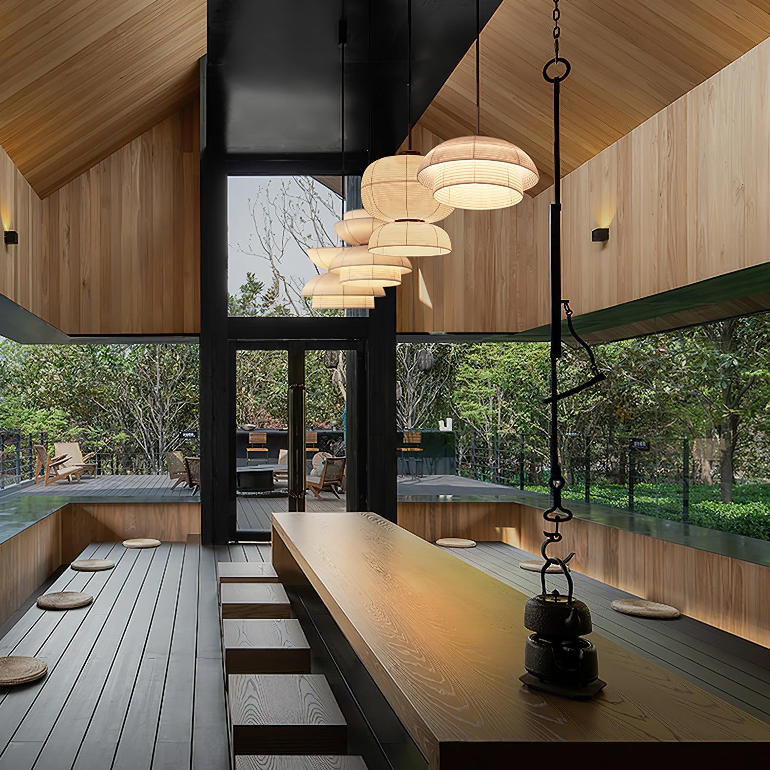 Varied fabric pendants in a row above a long wooden table and benches under a pitched wood roof beside garden glazing
