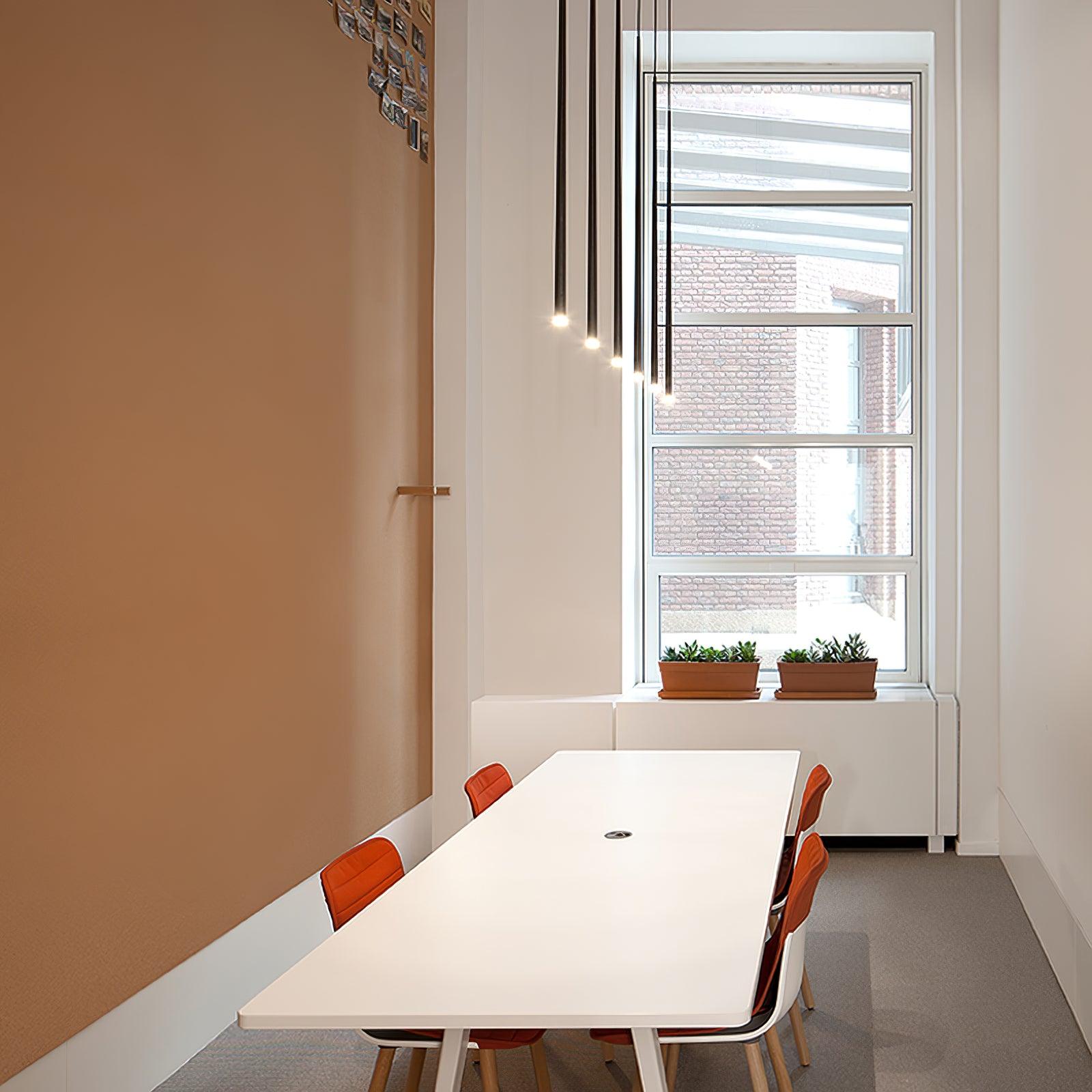 Illuminated black tube pendants above a long white table and orange chairs beside a tall window