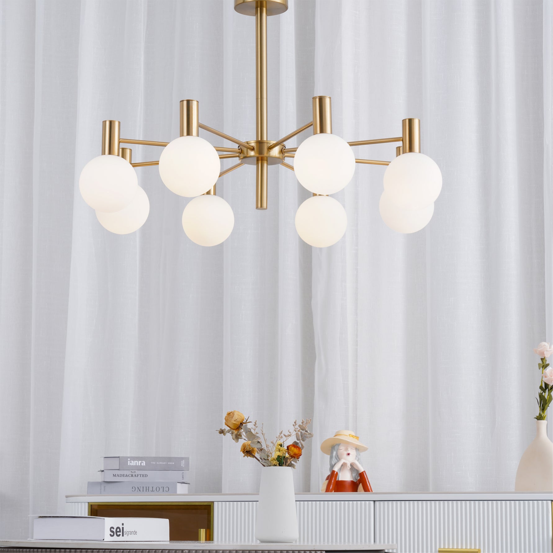 Lit white-globe chandelier in front of white sheers, above a ribbed cabinet with books, flowers and a hat-wearing figurine.