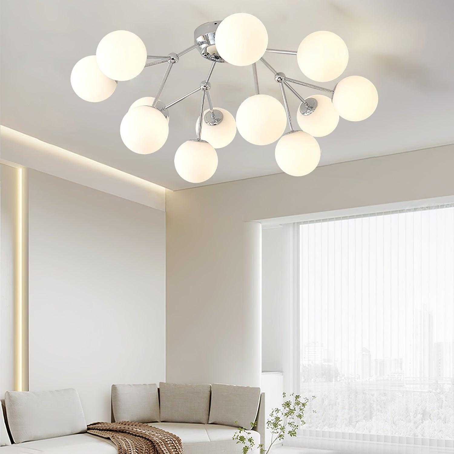Angled chrome globe cluster above a curved pale sofa with brown throw near sheer curtains and green branches