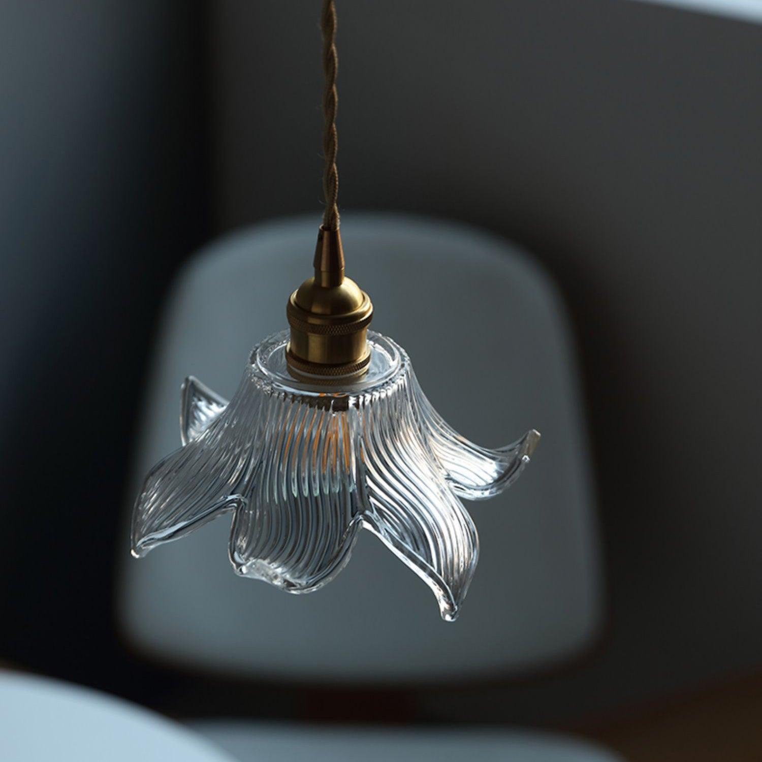 Angled clear petal shade and brass-toned upper fitting before a pale upholstered chair and dark grey wall.