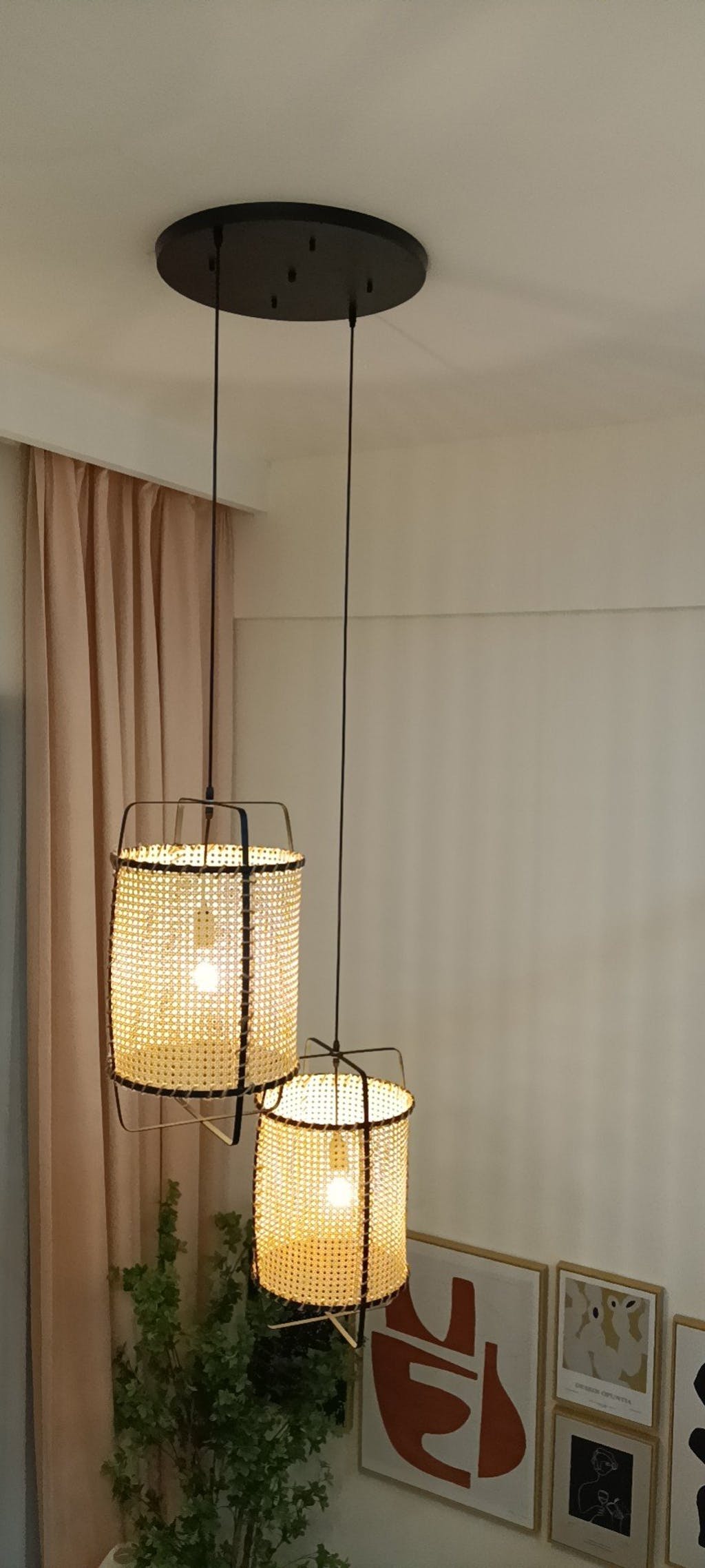 Two glowing cane pendants at different heights beneath a large shared black canopy near curtains and artwork