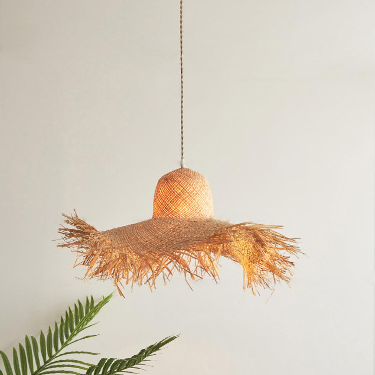 Warm-lit woven hat pendant against a pale wall with palm fronds beneath the loose fringed brim