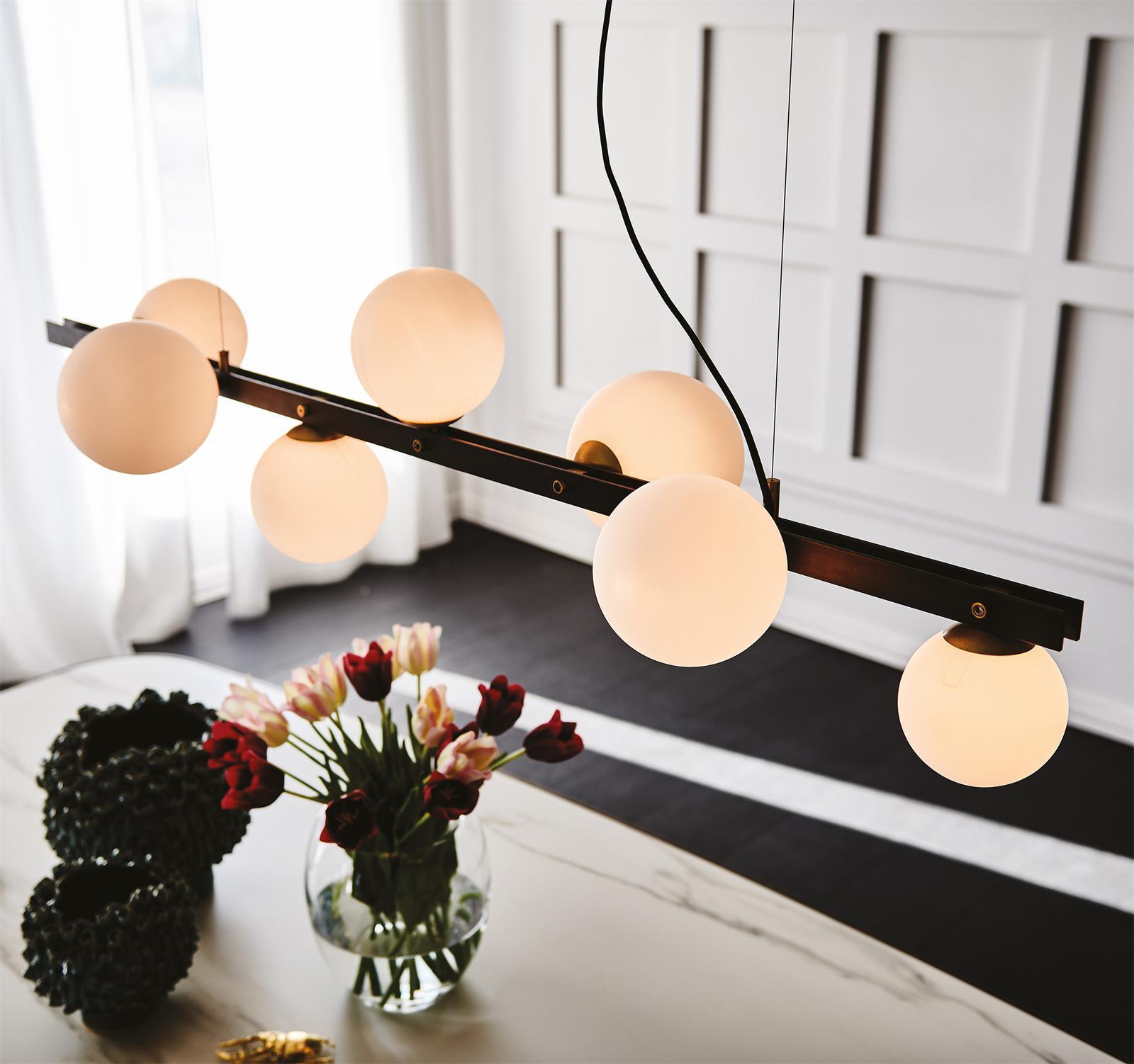 High diagonal view of seven glowing globes along black bar, fine support lines and curved cord, above tulips and green bowls.