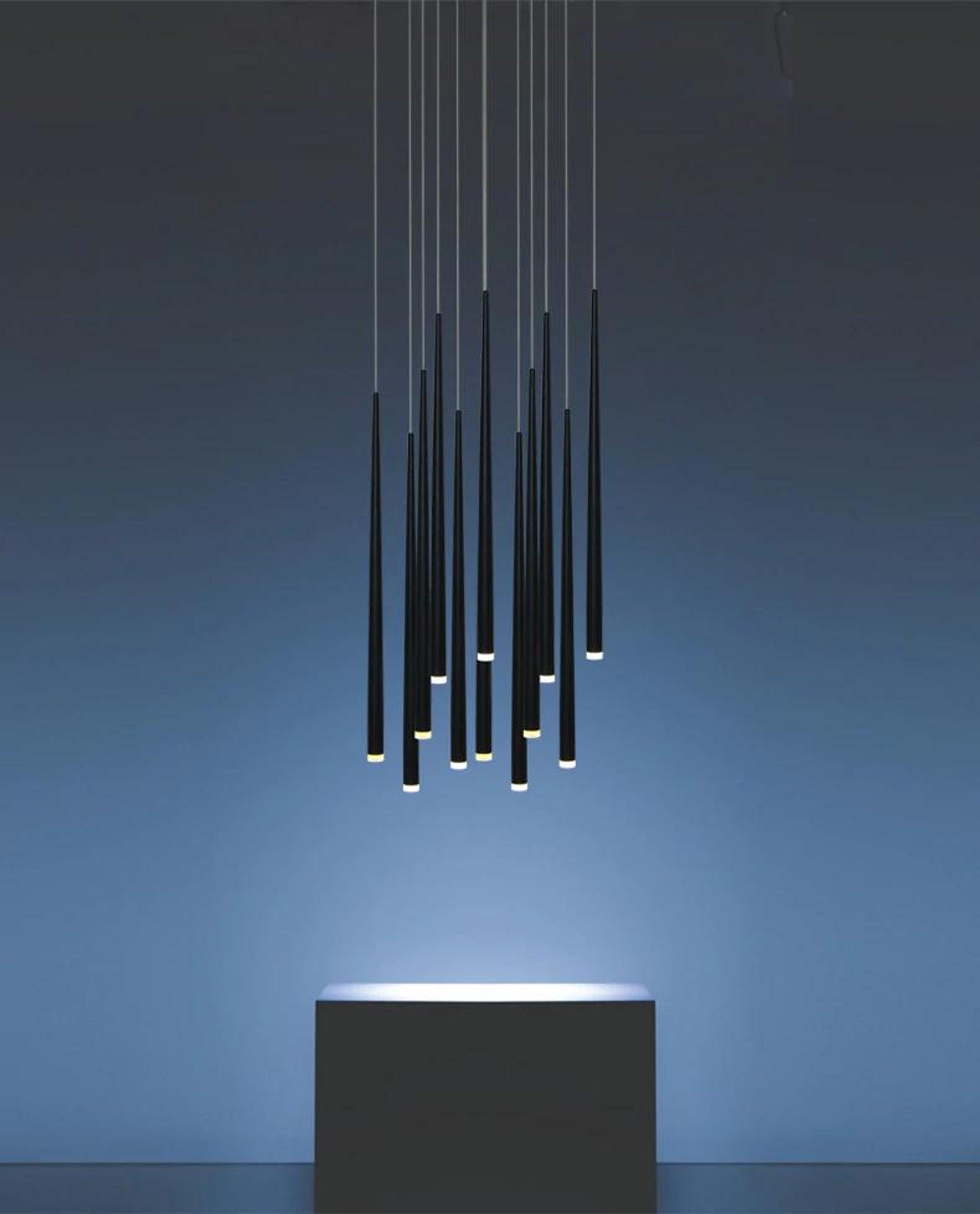 Black tube pendant cluster illuminated against a blue wall above a black display plinth