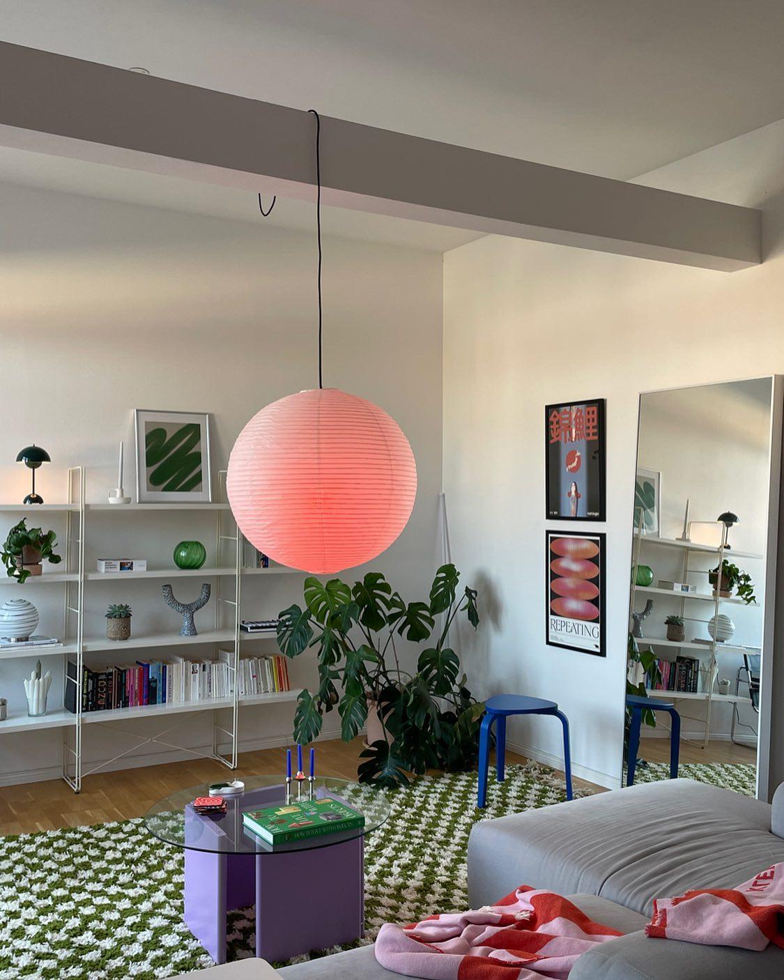 Pink-looking round illuminated lantern on black cord beside grey sofa, shelves, plant and large mirror