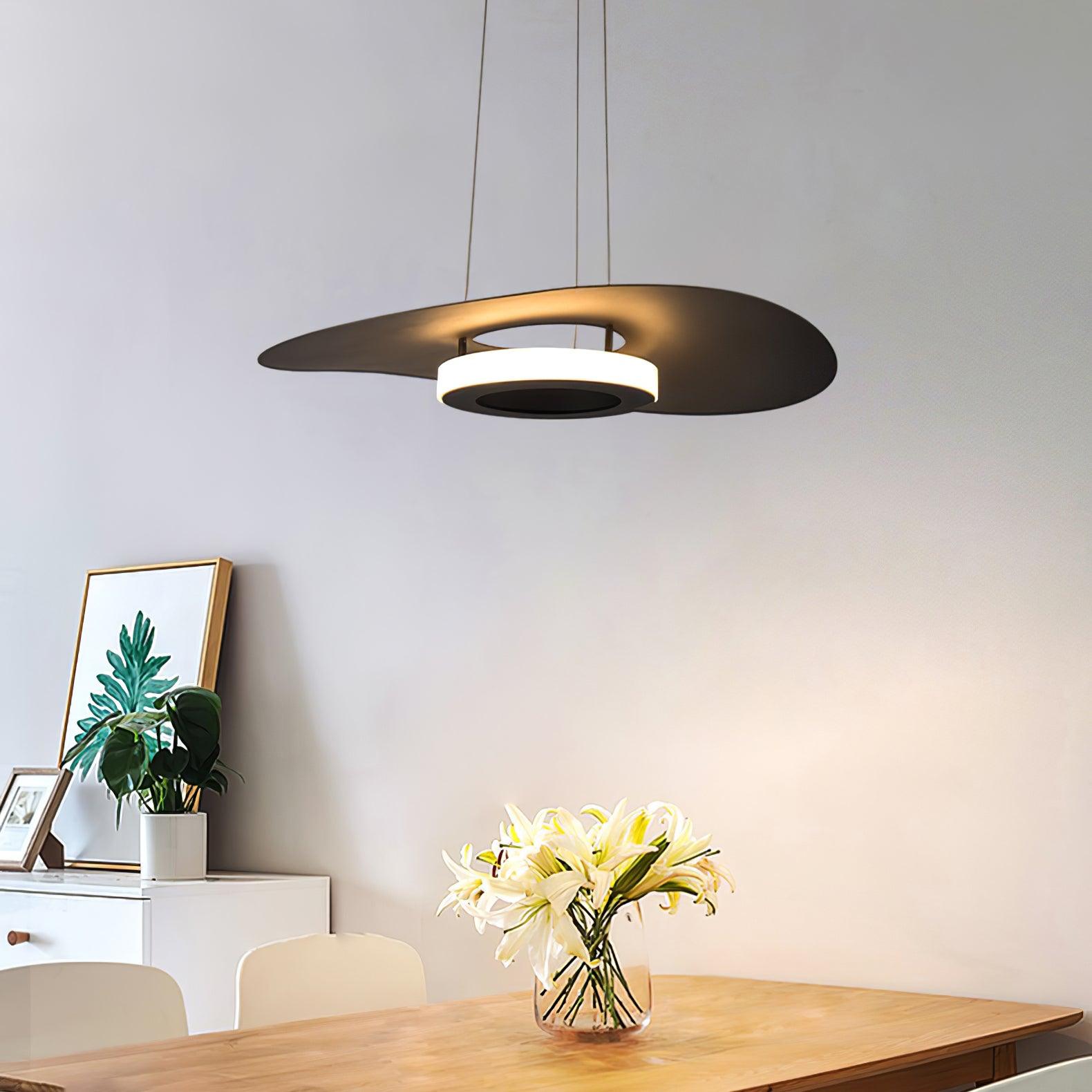 Dark disc pendant and glowing lower ring above wooden dining table with lilies, leaf picture and white cabinet