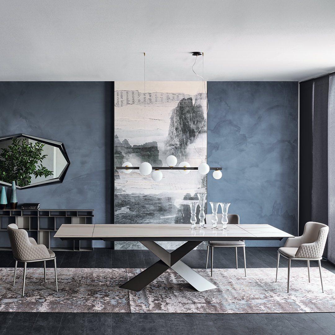 Wide globe pendant above pale crossed-base table and clear vases, against blue-grey walls, monochrome mural and polygon mirror.