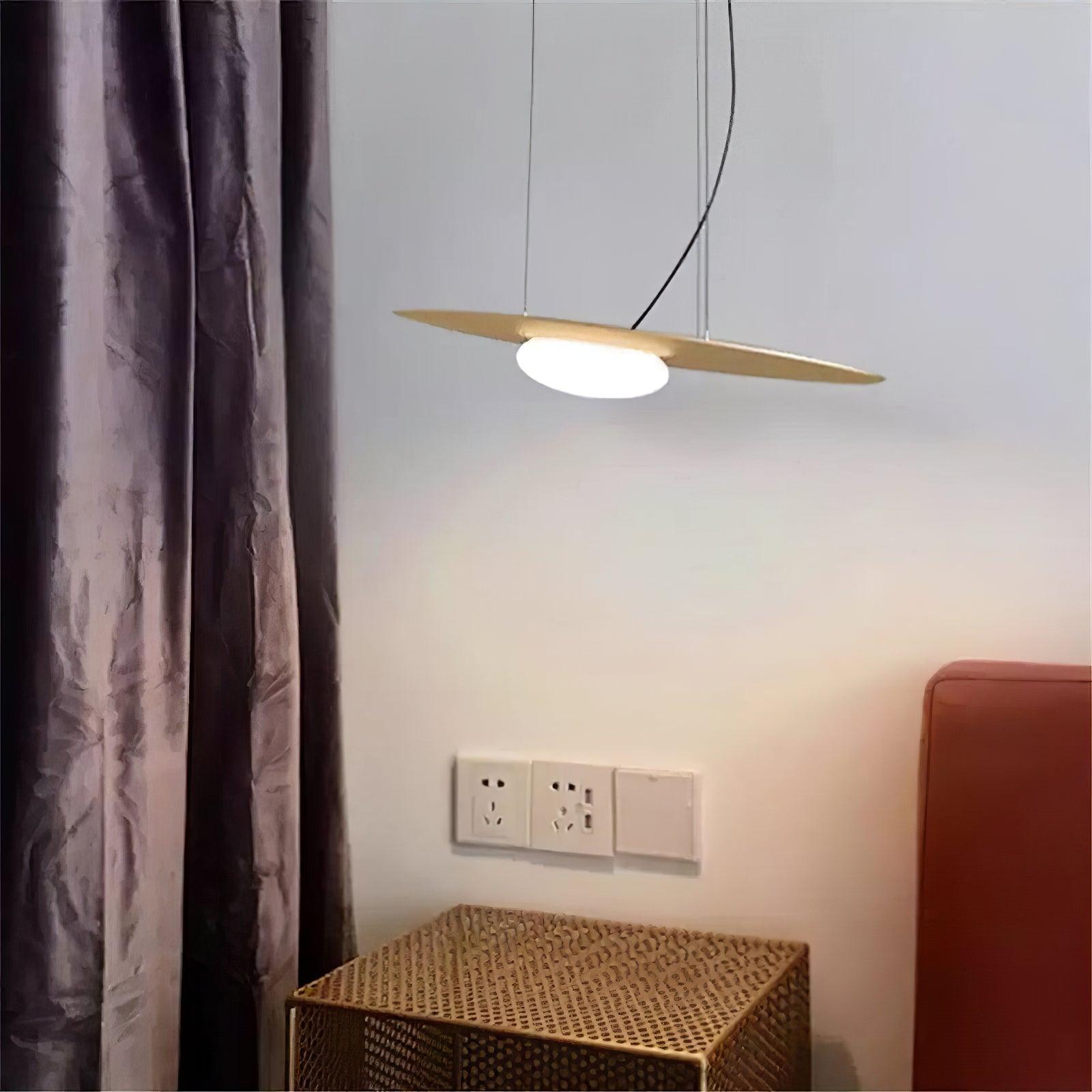 Close view of gold-tone glowing disc beside dark curtains and rust headboard, above woven bedside cabinet and wall sockets.