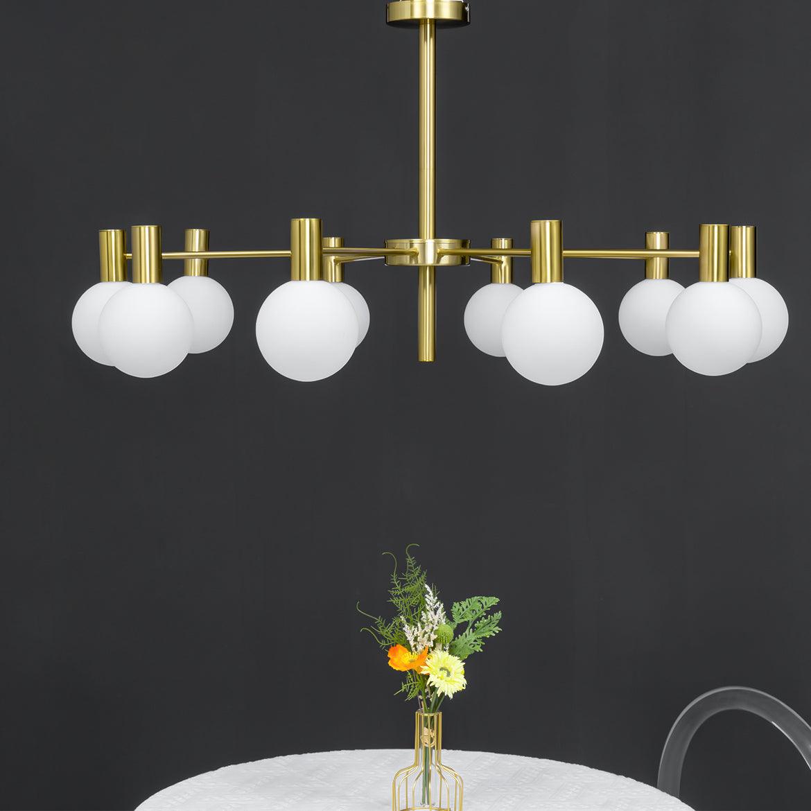 Unlit white-globe chandelier with gold-tone arms and rod above the edge of a round table and a vase of flowers.